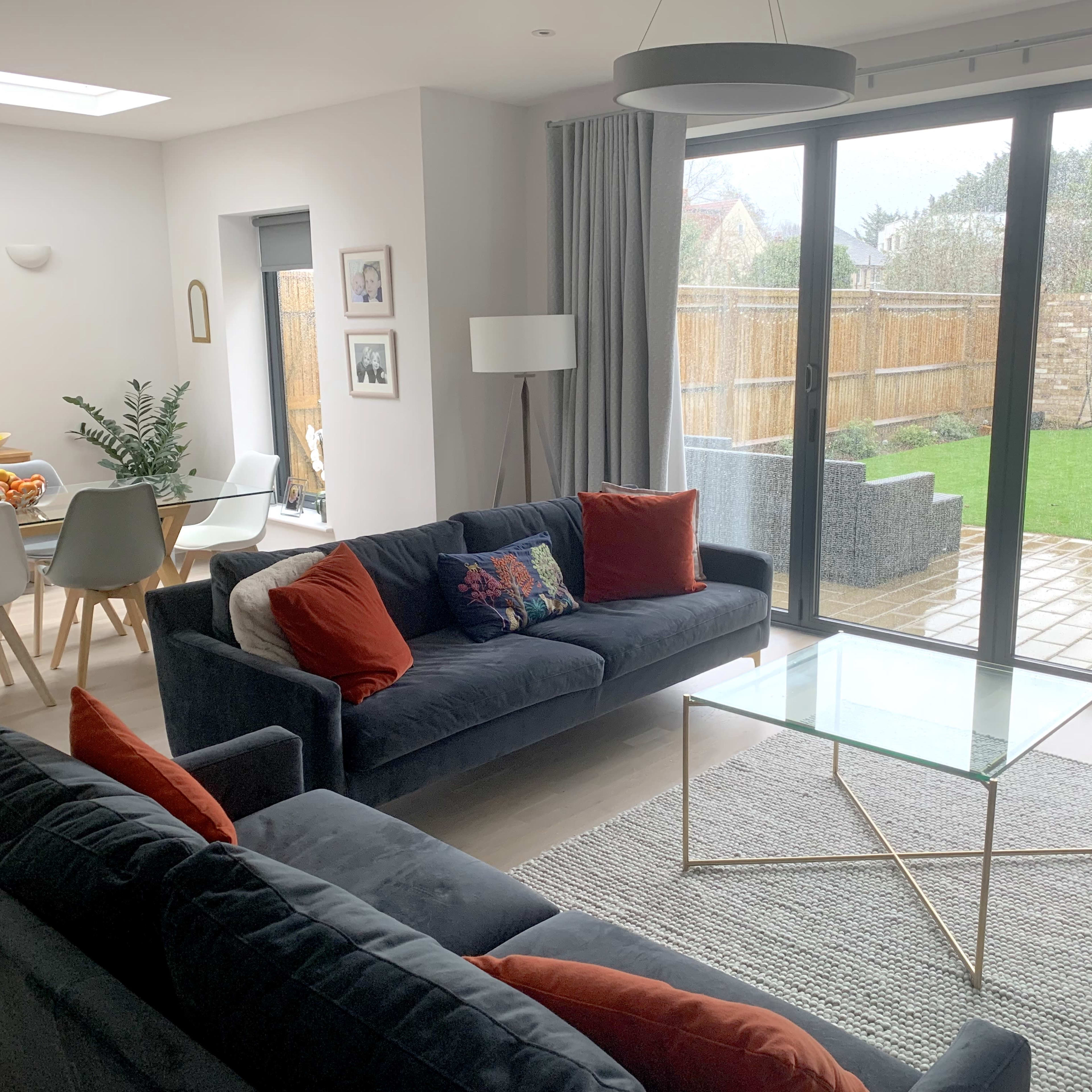 A modern living room with two sofas facing a glass coffee table, large windows overlooking a backyard, and a dining area in the background.