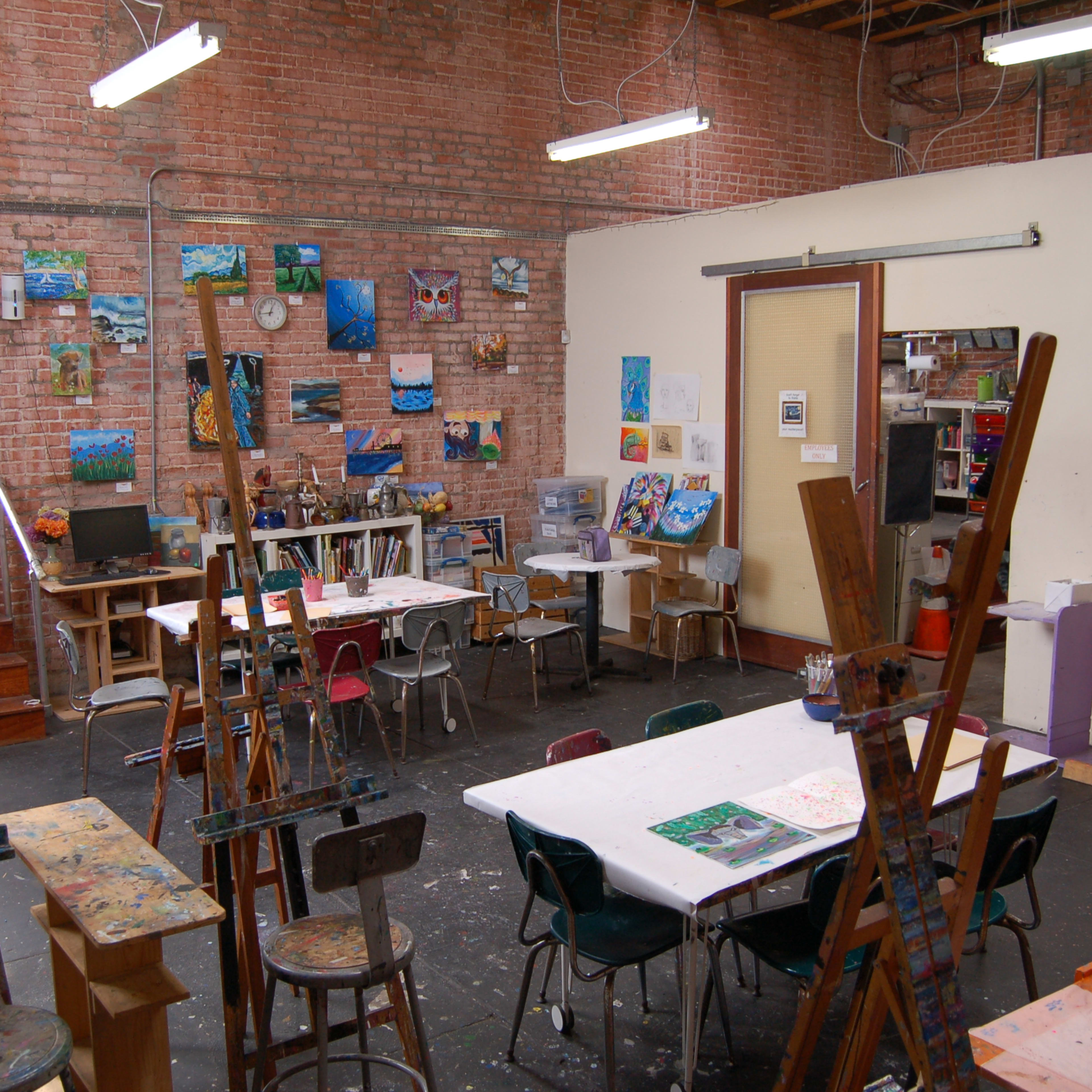 An art studio features easels and tables surrounded by paintings on the walls, with an entrance visible in the background.