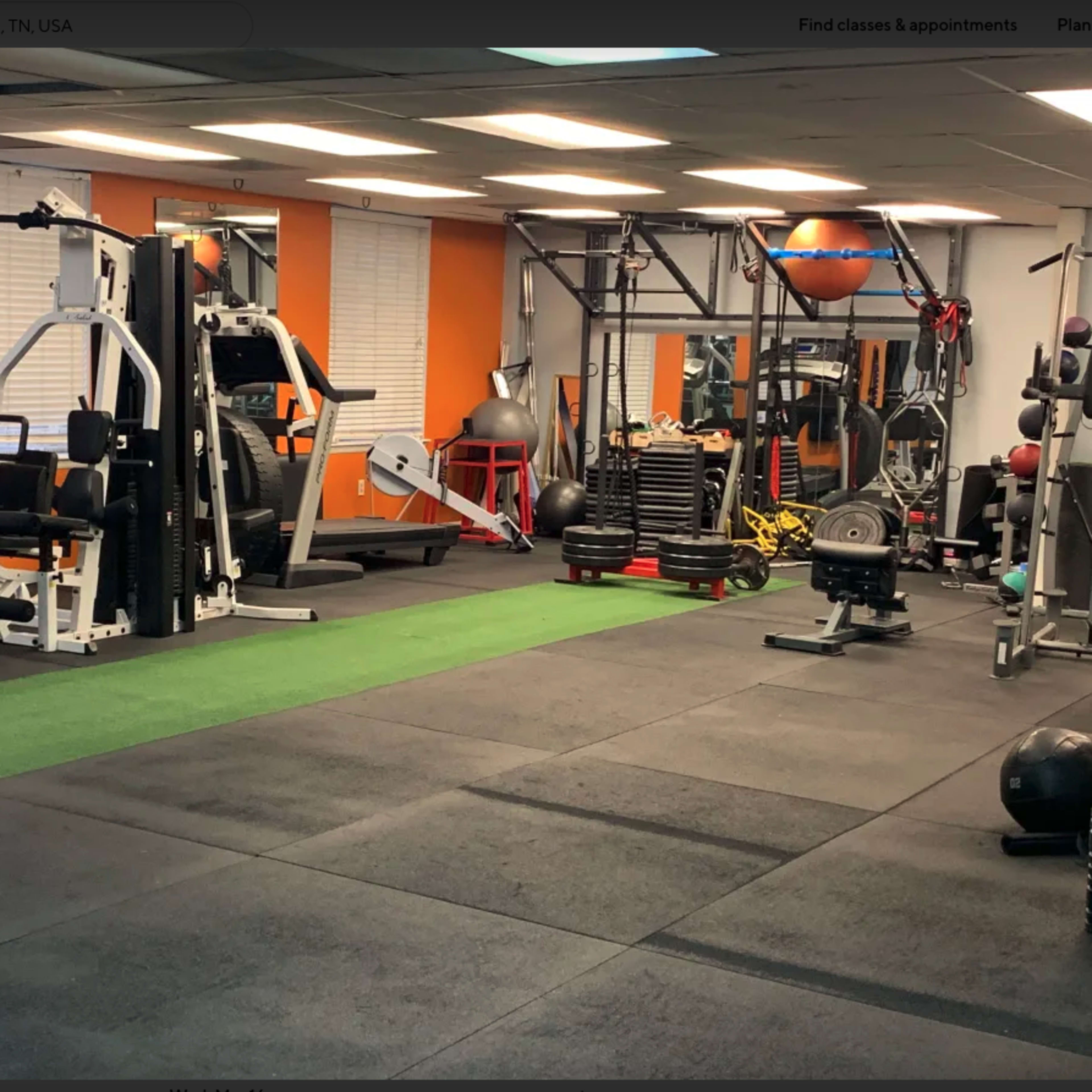 A fitness gym equipped with various exercise machines and weights, featuring a green turf area and black rubber flooring.