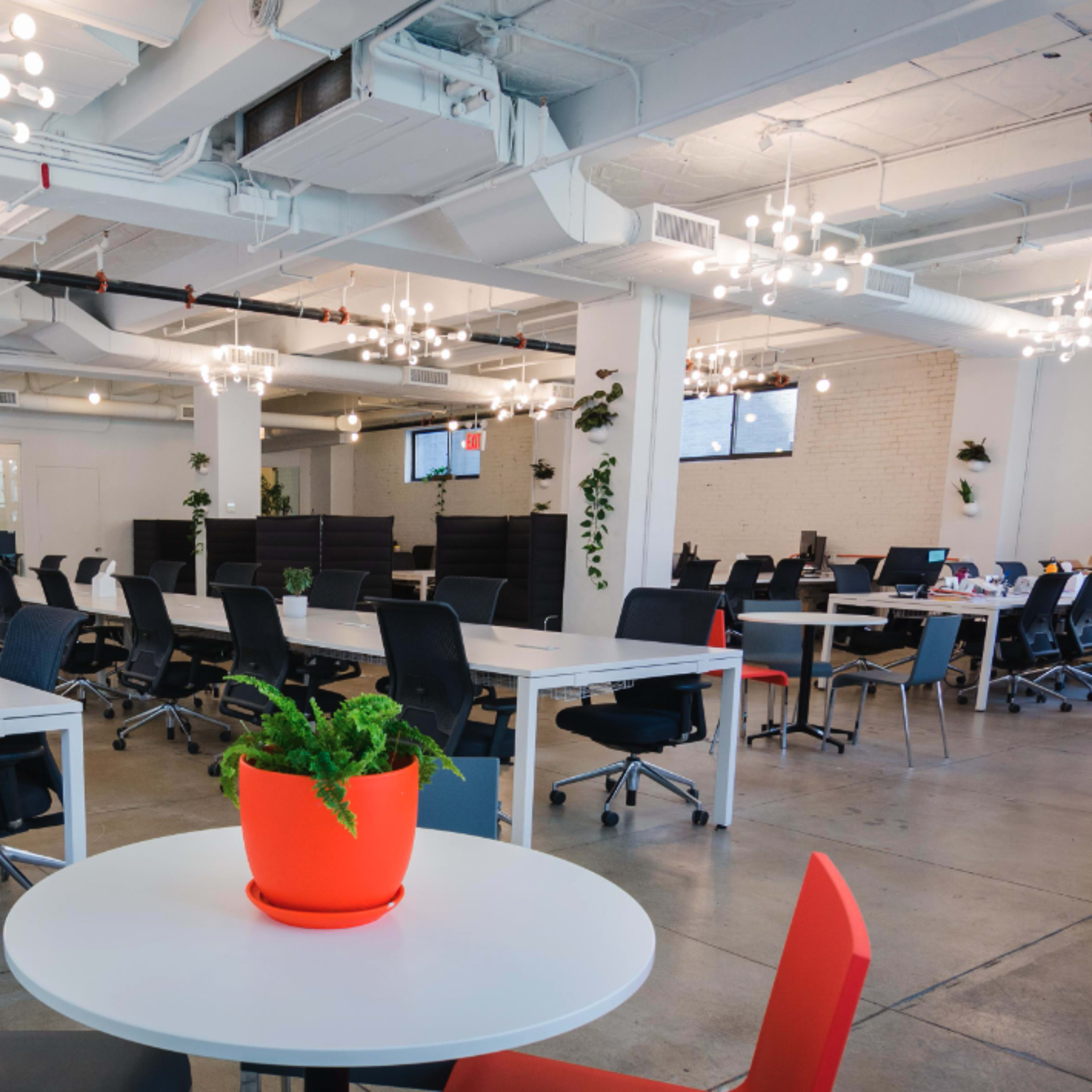 A spacious, modern office features long white tables, black chairs, and scattered greenery, with bright lighting and a few workstations visible in the background.