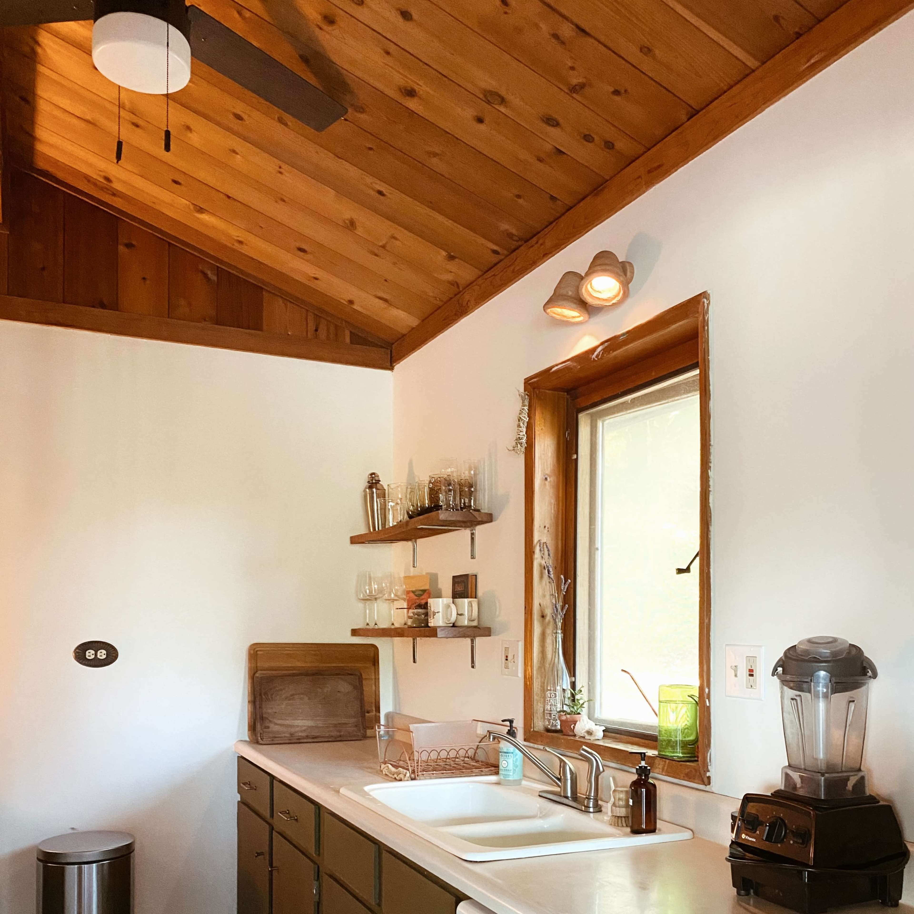 A kitchen features wooden ceiling beams, beige walls, and green cabinetry, with a window above the sink and appliances on the counter.