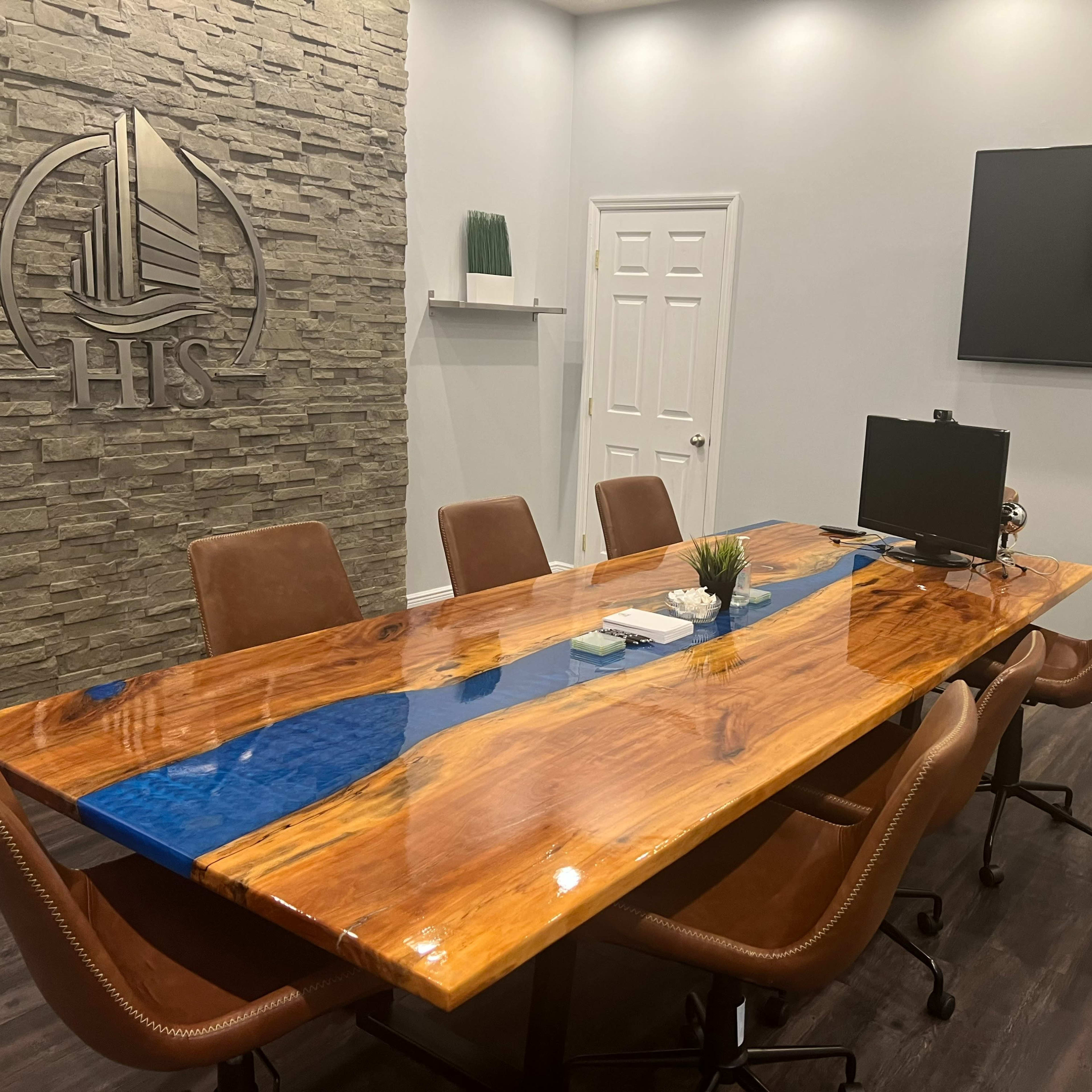 A modern conference room with a large wooden table featuring a blue resin river, surrounded by brown chairs, and equipped with a wall-mounted television and a decorative stone wall.