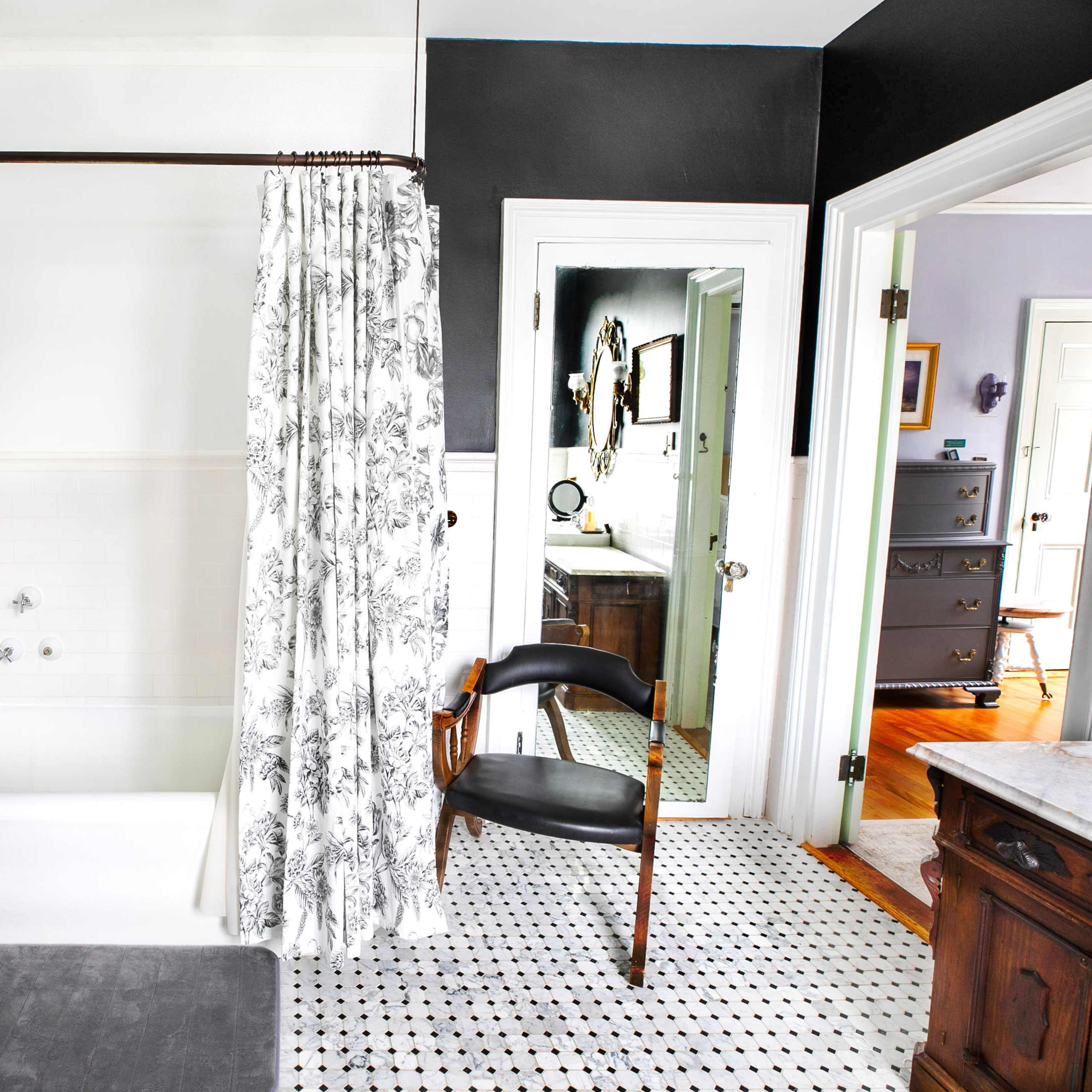 A bathroom featuring a bathtub with a shower curtain, a toilet, a chair, and a view of an adjoining room through an open door.