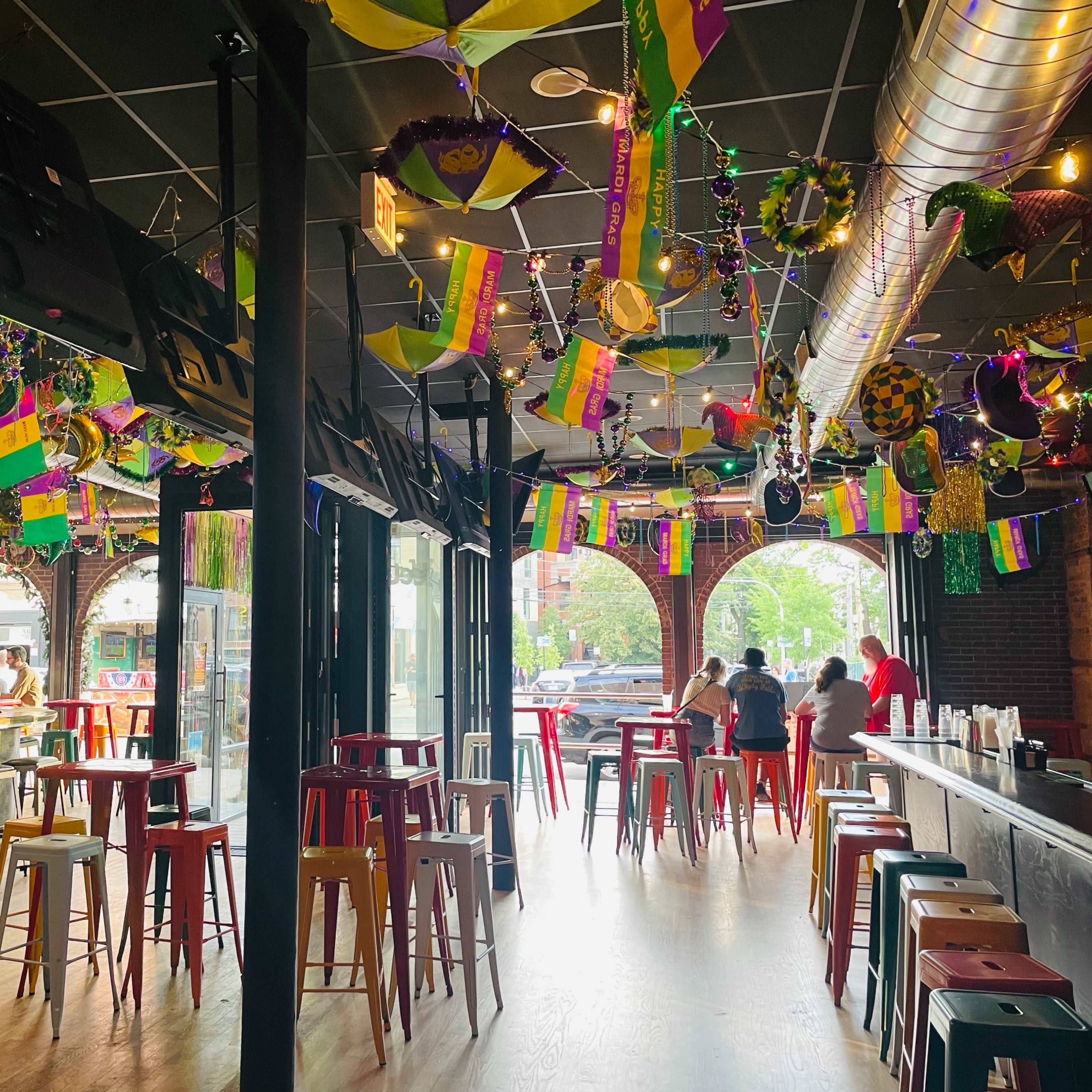 The interior of a bar features colorful Mardi Gras decorations, including beads and banners, with high tables and stools arranged throughout the space.