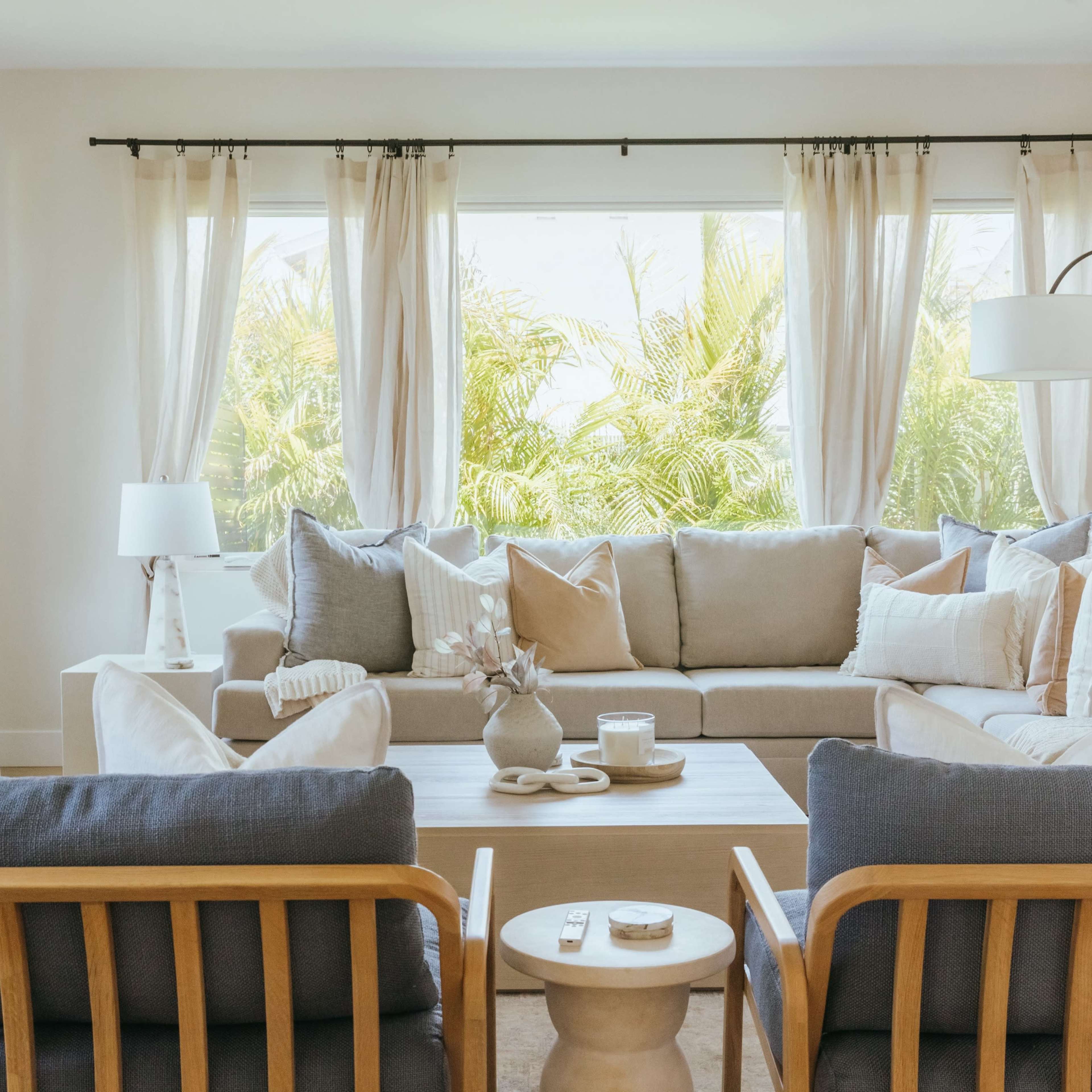 A bright living room features a light-colored sofa surrounded by armchairs, coffee table, and large windows with sheer curtains offering a view of greenery.