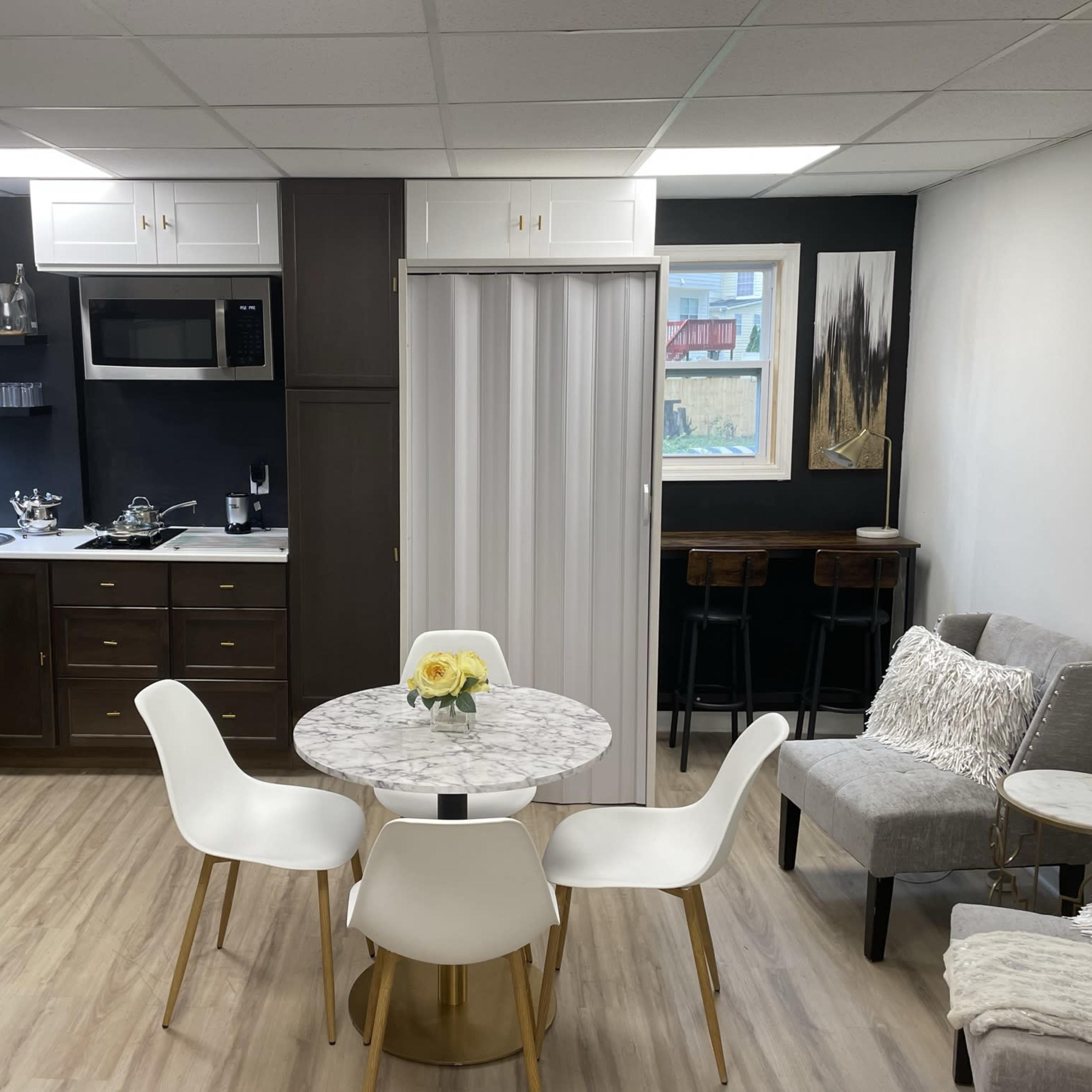 A modern kitchen and dining area featuring a round marble table with white chairs, stainless steel appliances, and a gray sofa against a dark wall.