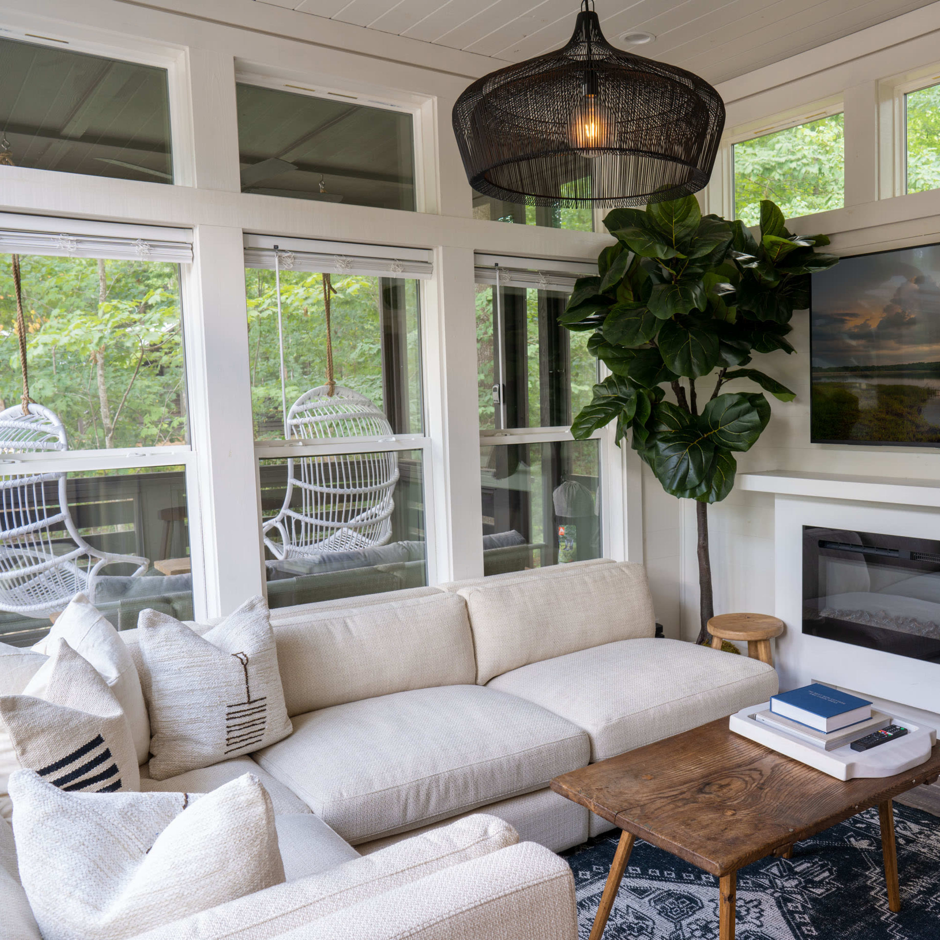 A cozy living room features a white sectional sofa, a wooden coffee table, and a mounted TV above a sleek fireplace, surrounded by large windows and greenery.