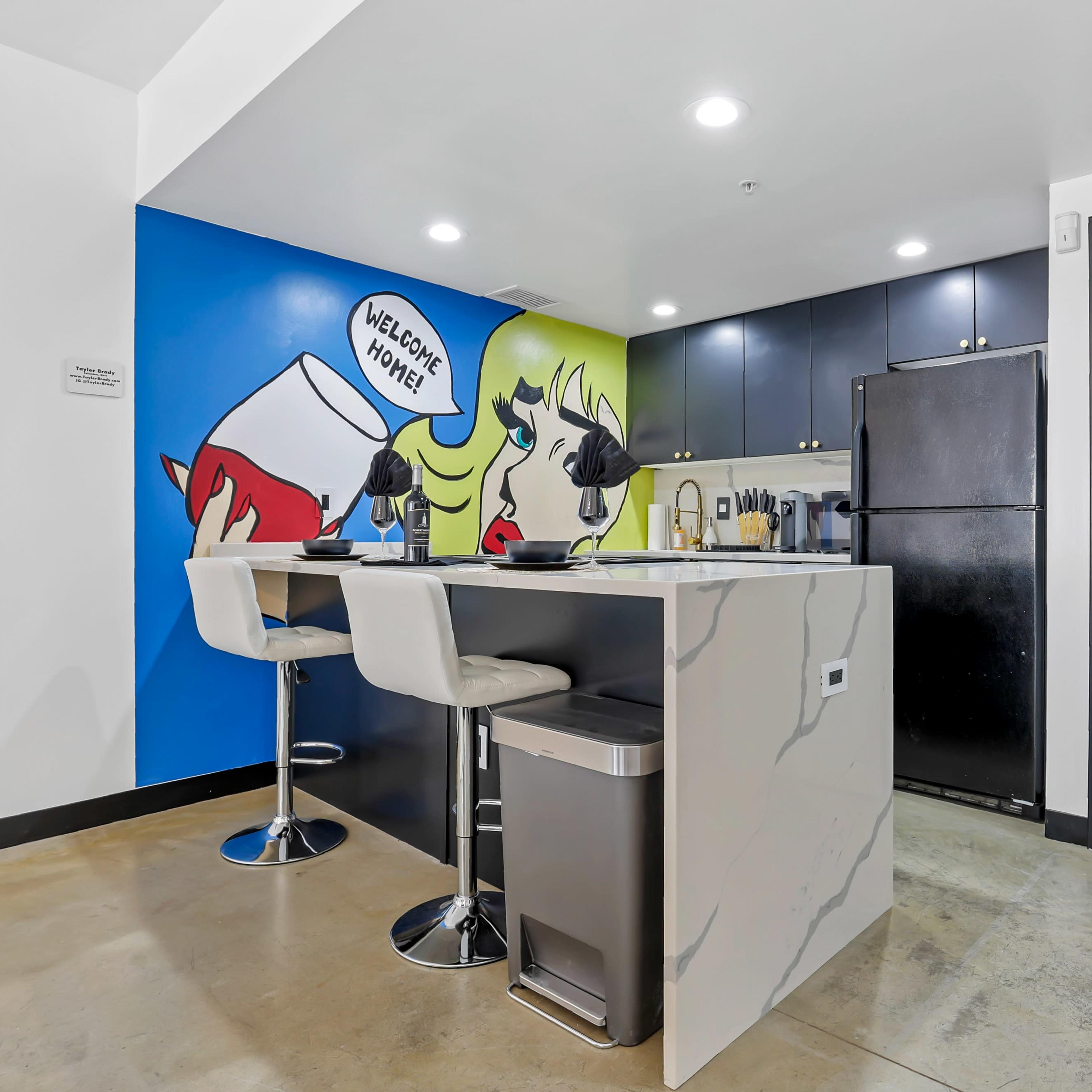 A modern kitchen features a bar with two stools and a colorful wall mural, welcoming guests.