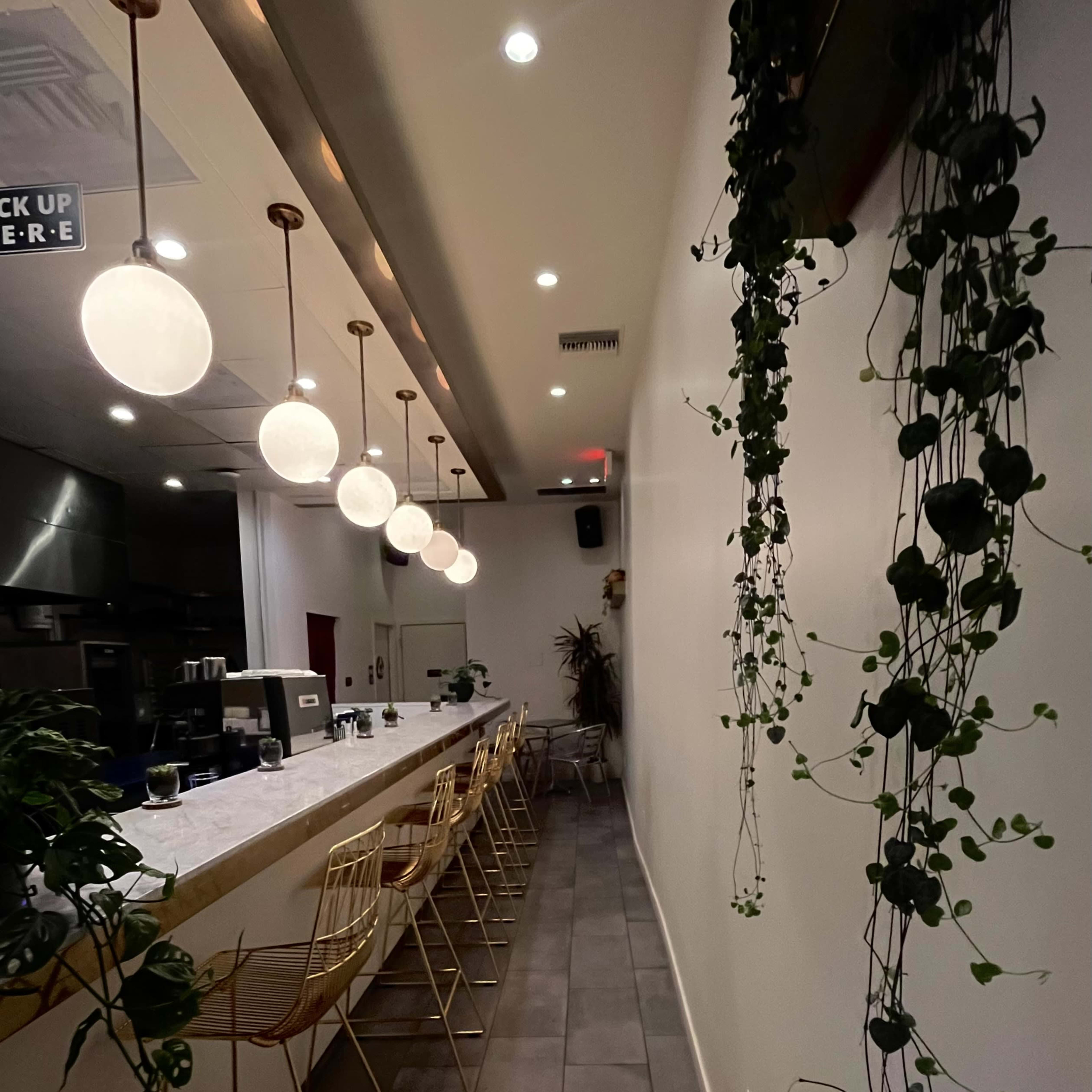 A narrow, modern bar area with a long countertop, golden bar stools, hanging pendant lights, and plants trailing down from the ceiling.