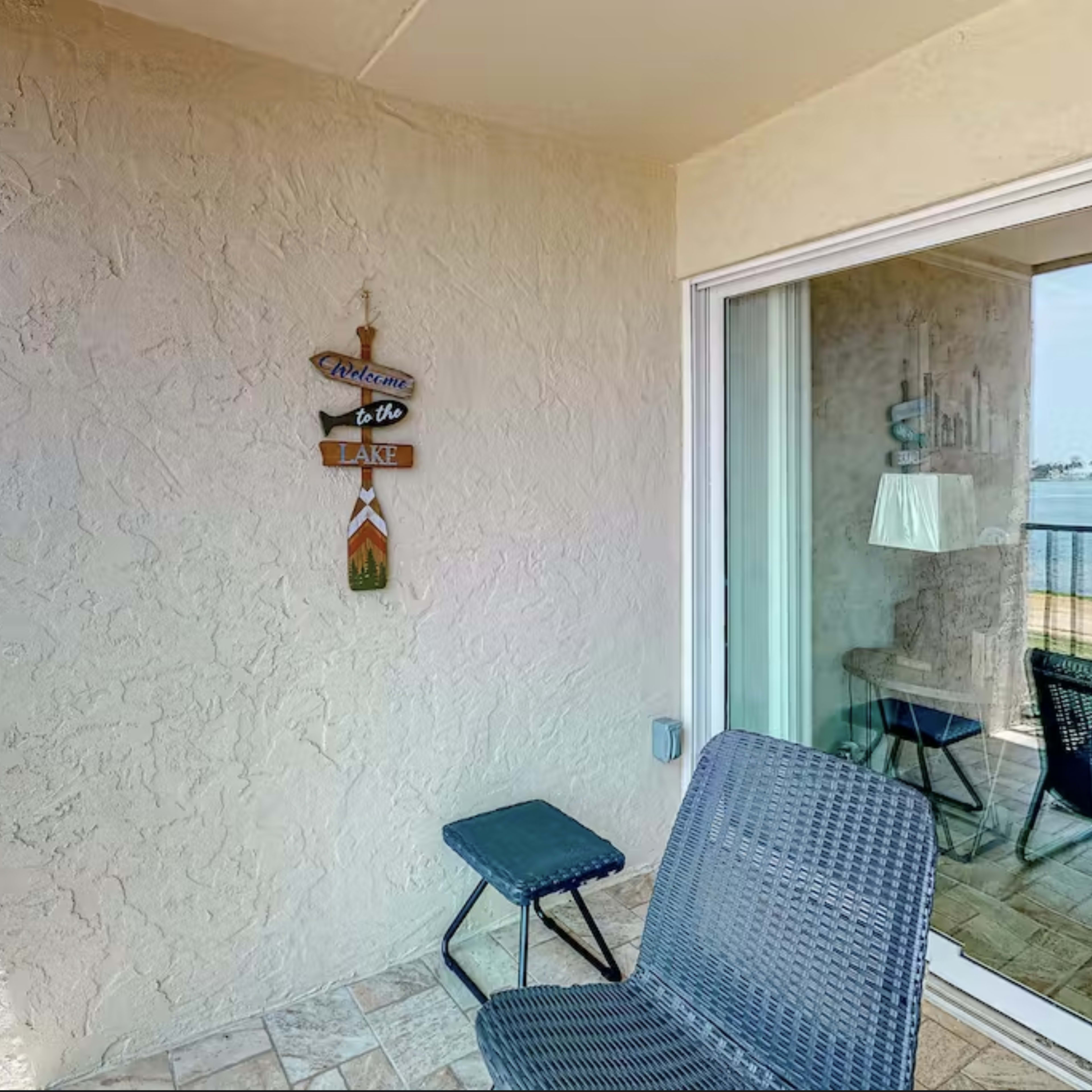 A balcony features a textured wall with a "Welcome to the Lake" sign and a chair beside a small table.