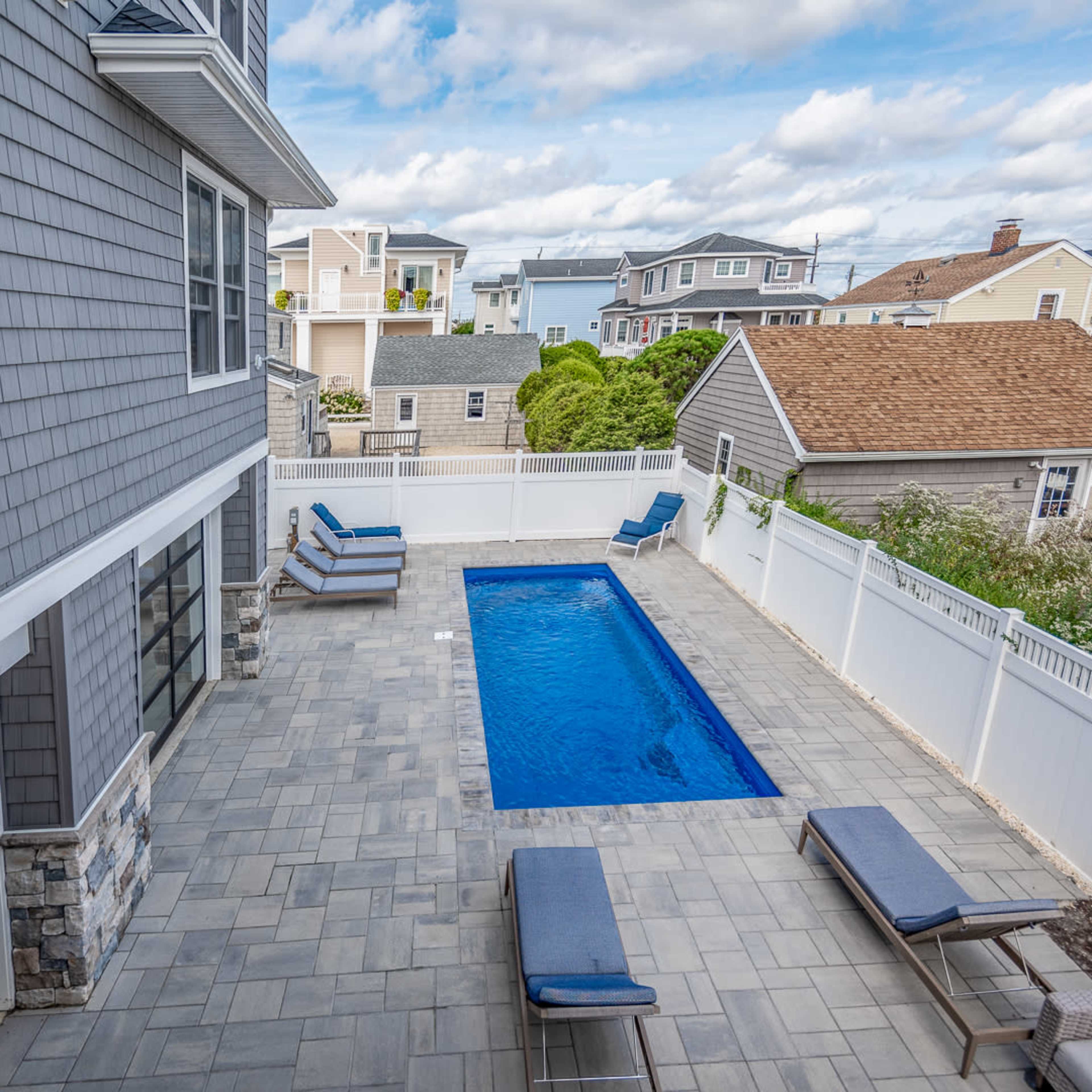 A rectangular swimming pool surrounded by a paved patio area and two lounge chairs, set against a backdrop of various residential buildings.