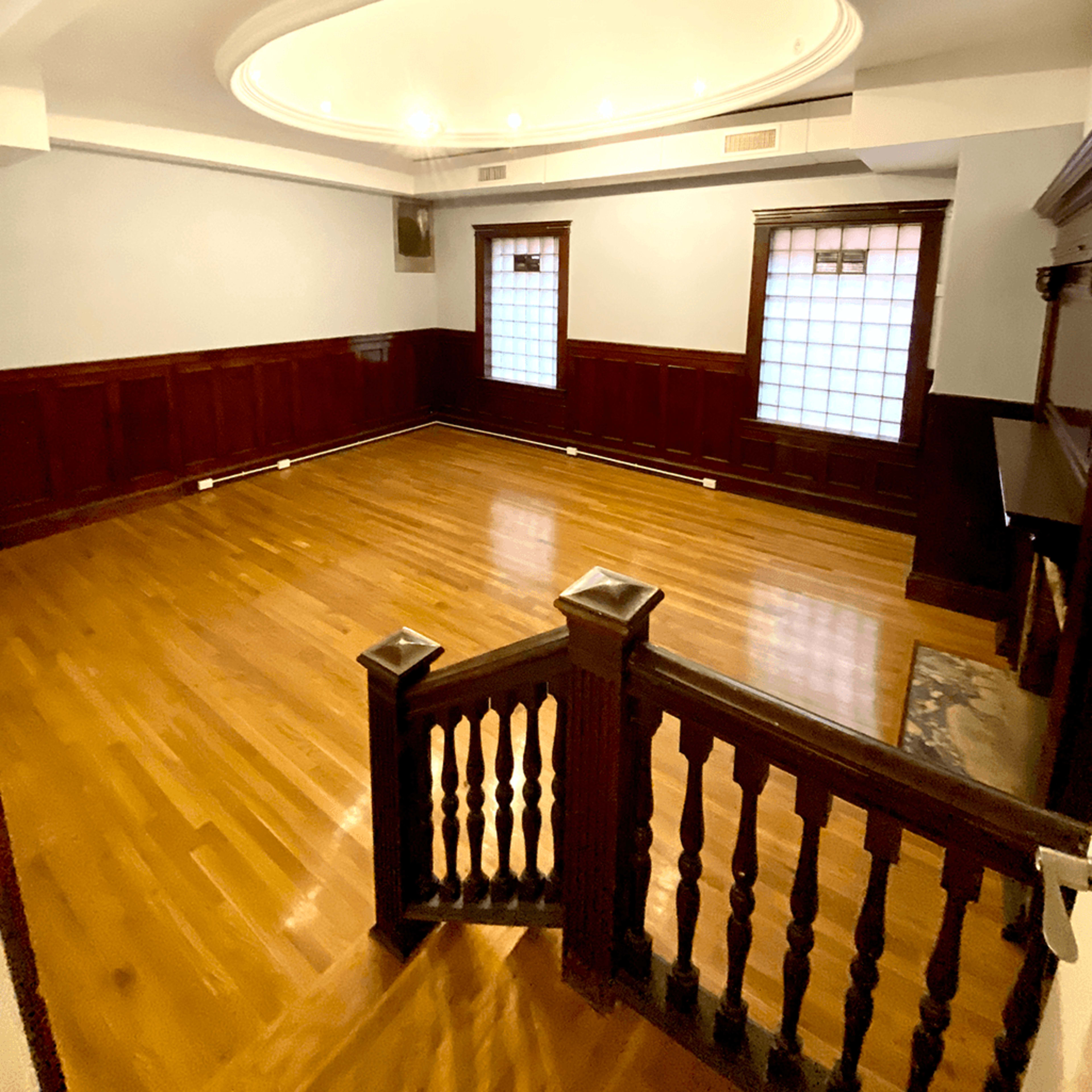 An empty room with wooden flooring, circular ceiling lights, and wainscoted walls, featuring two windows with grid patterns.