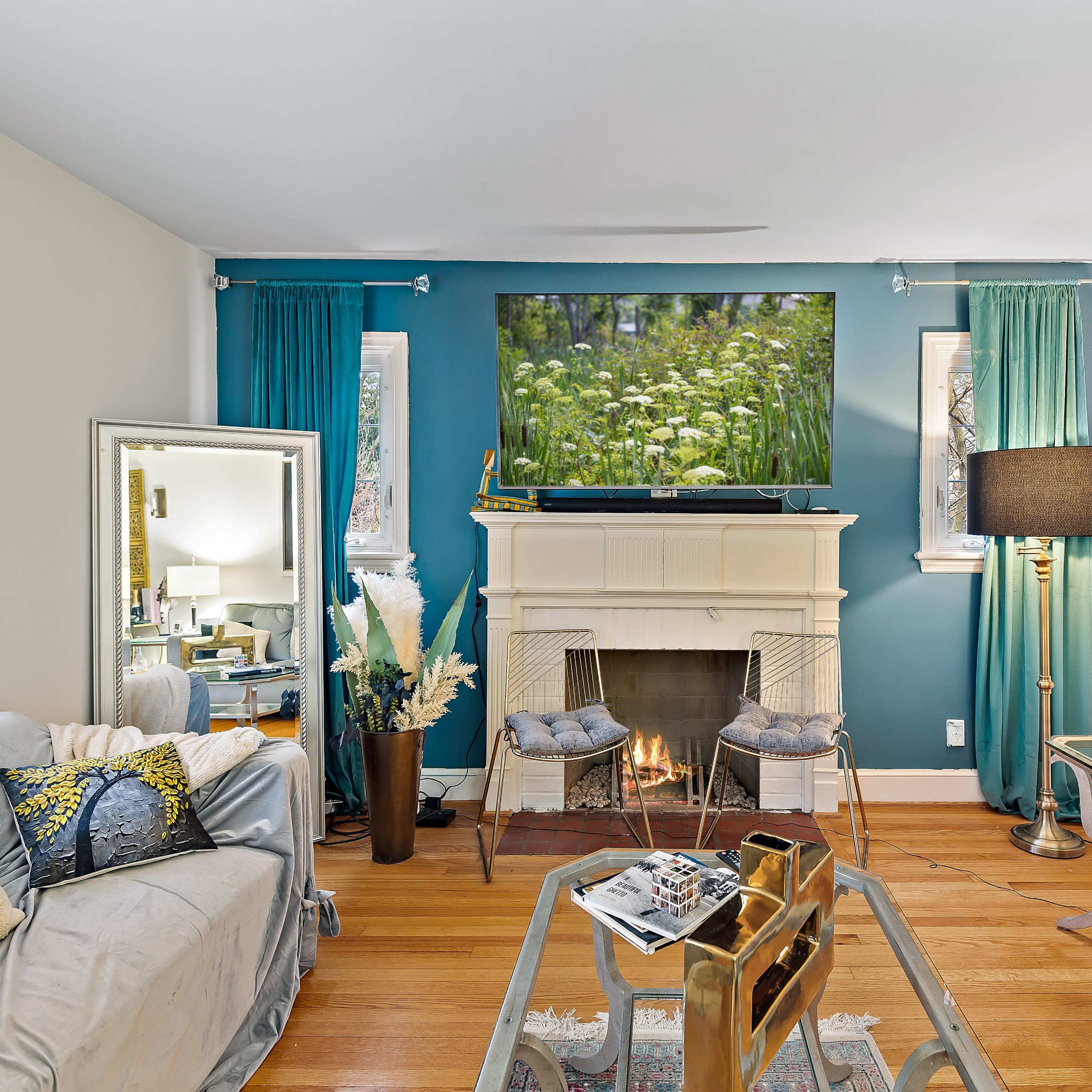 A living room features a television above a fireplace, surrounded by teal curtains, a coffee table, and various seating options.