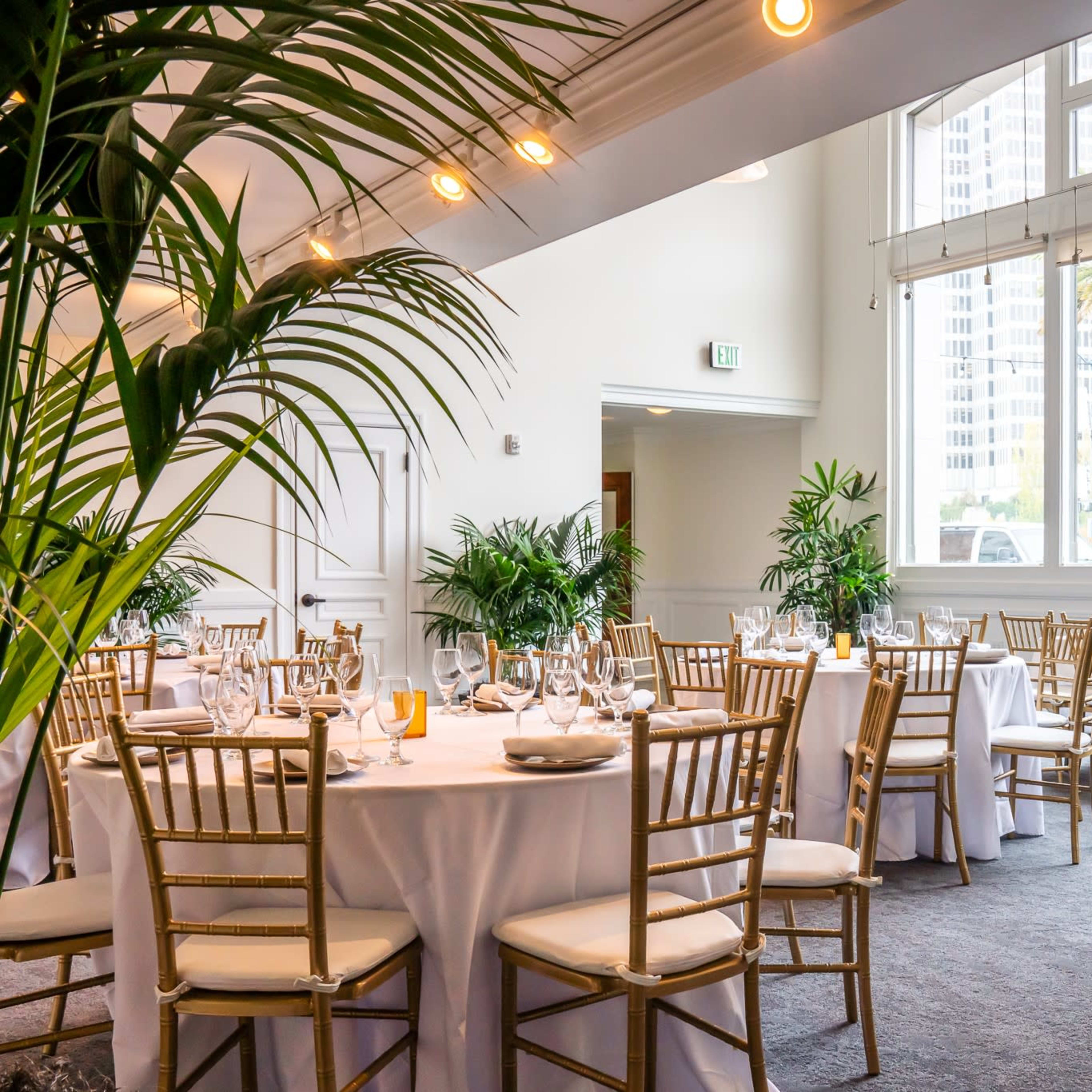 A banquet room with round tables dressed in white tablecloths and golden chairs, surrounded by large windows and indoor plants.