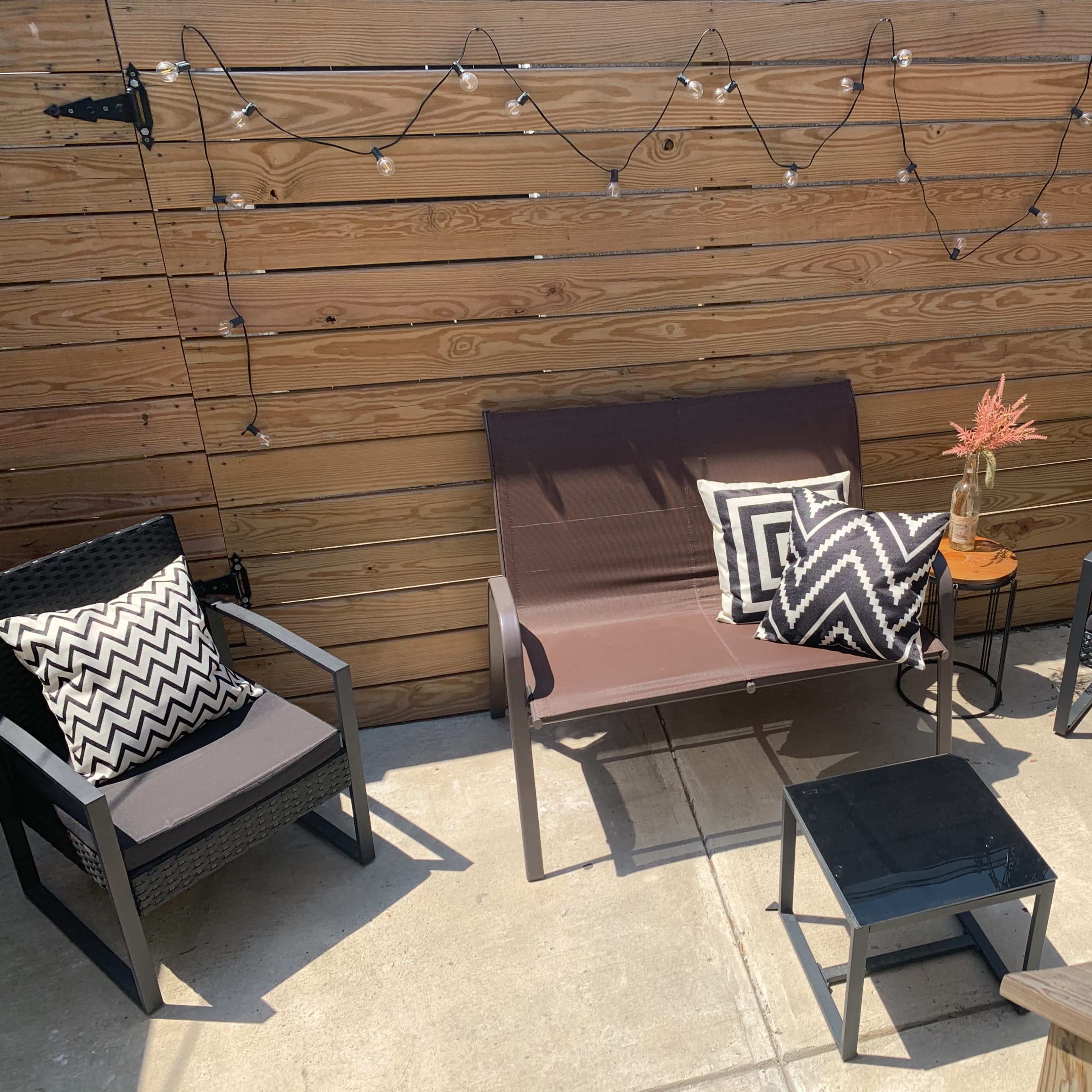A small outdoor seating area with a double chair, two single chairs with patterned cushions, and a small table, surrounded by wooden walls and string lights.