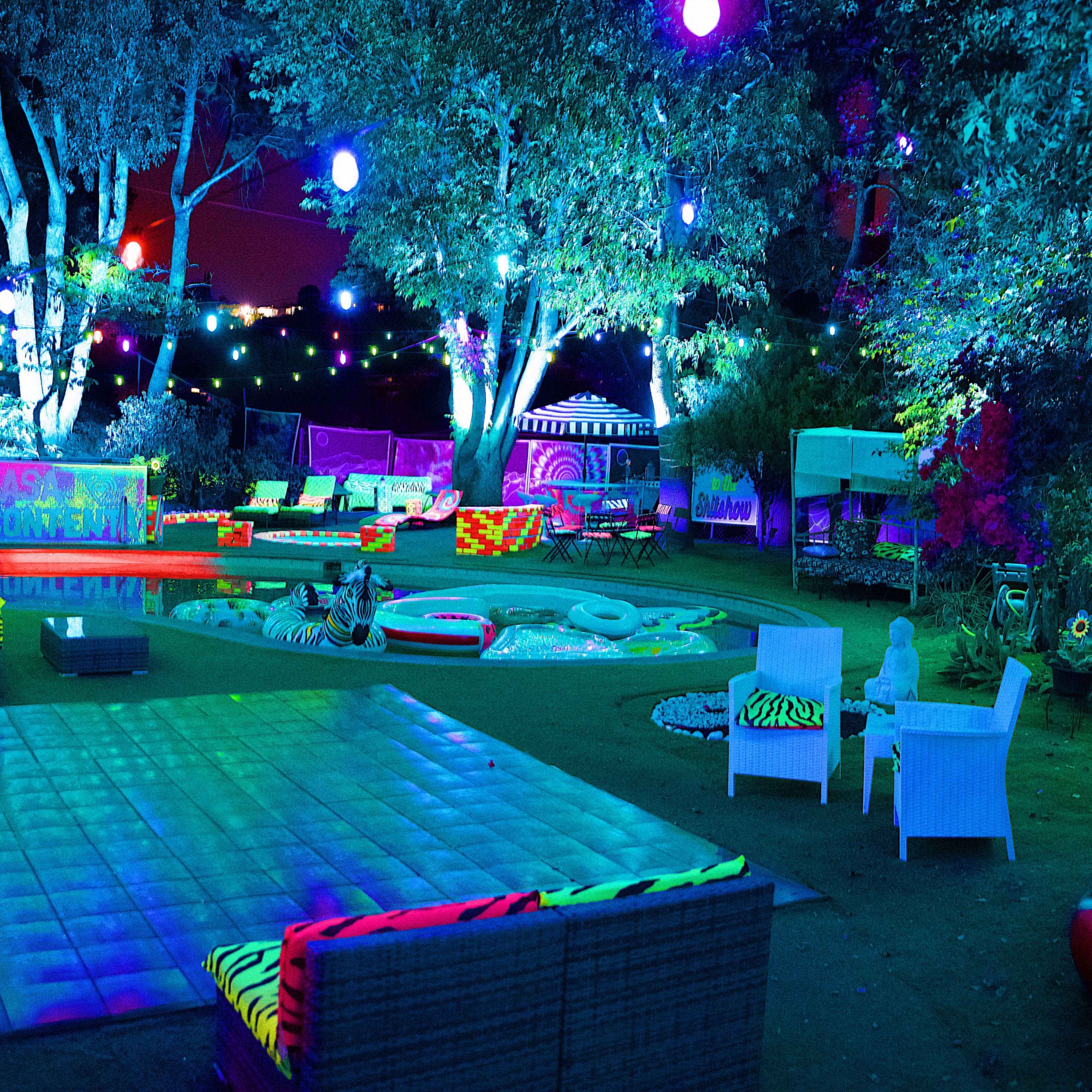 The scene shows a brightly lit outdoor party area with colorful lights, various seating arrangements, and vibrant decorations among trees and garden elements.