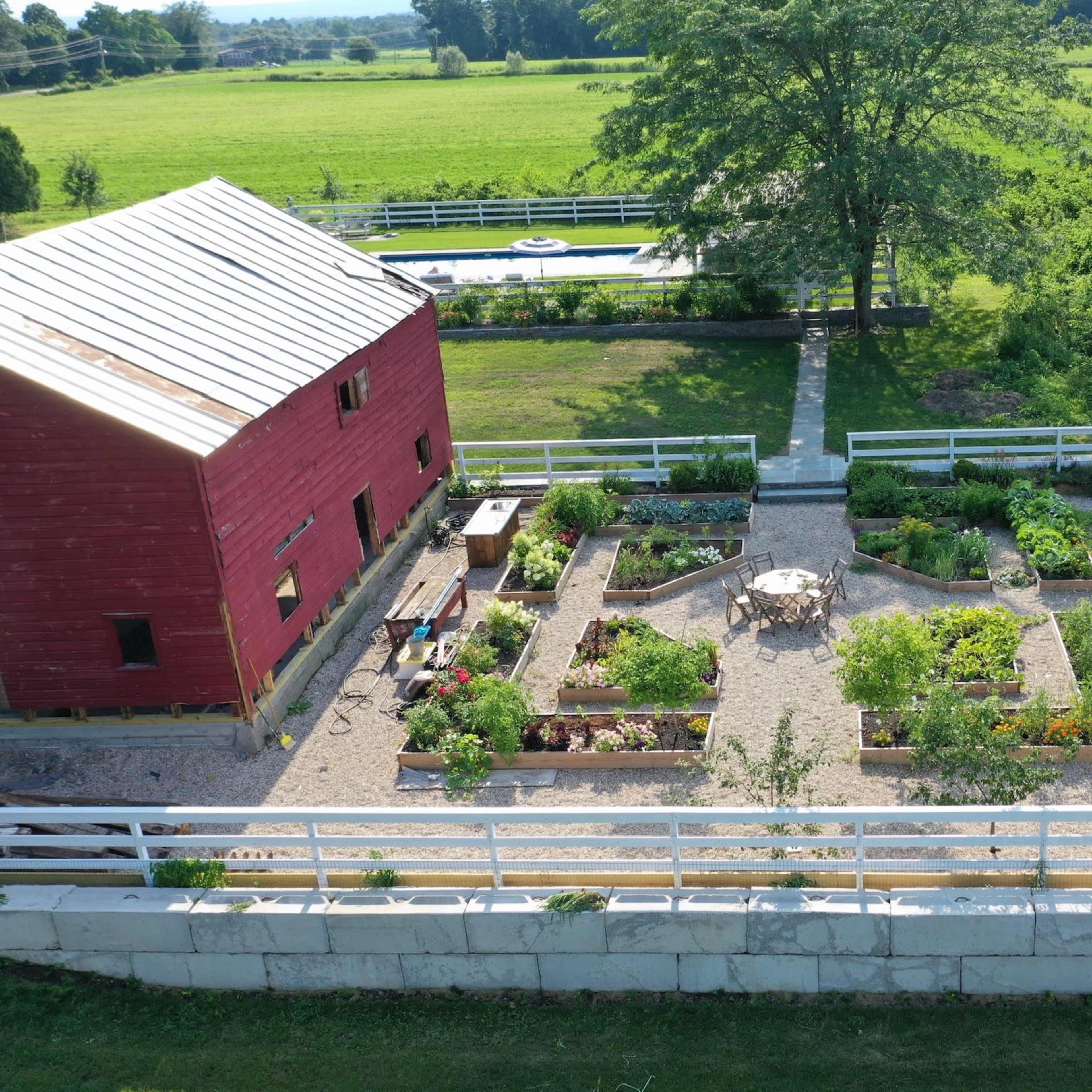 A large garden arranged in a geometric layout with various plants, bordered by a red barn and a grassy area, alongside a pool in the background.