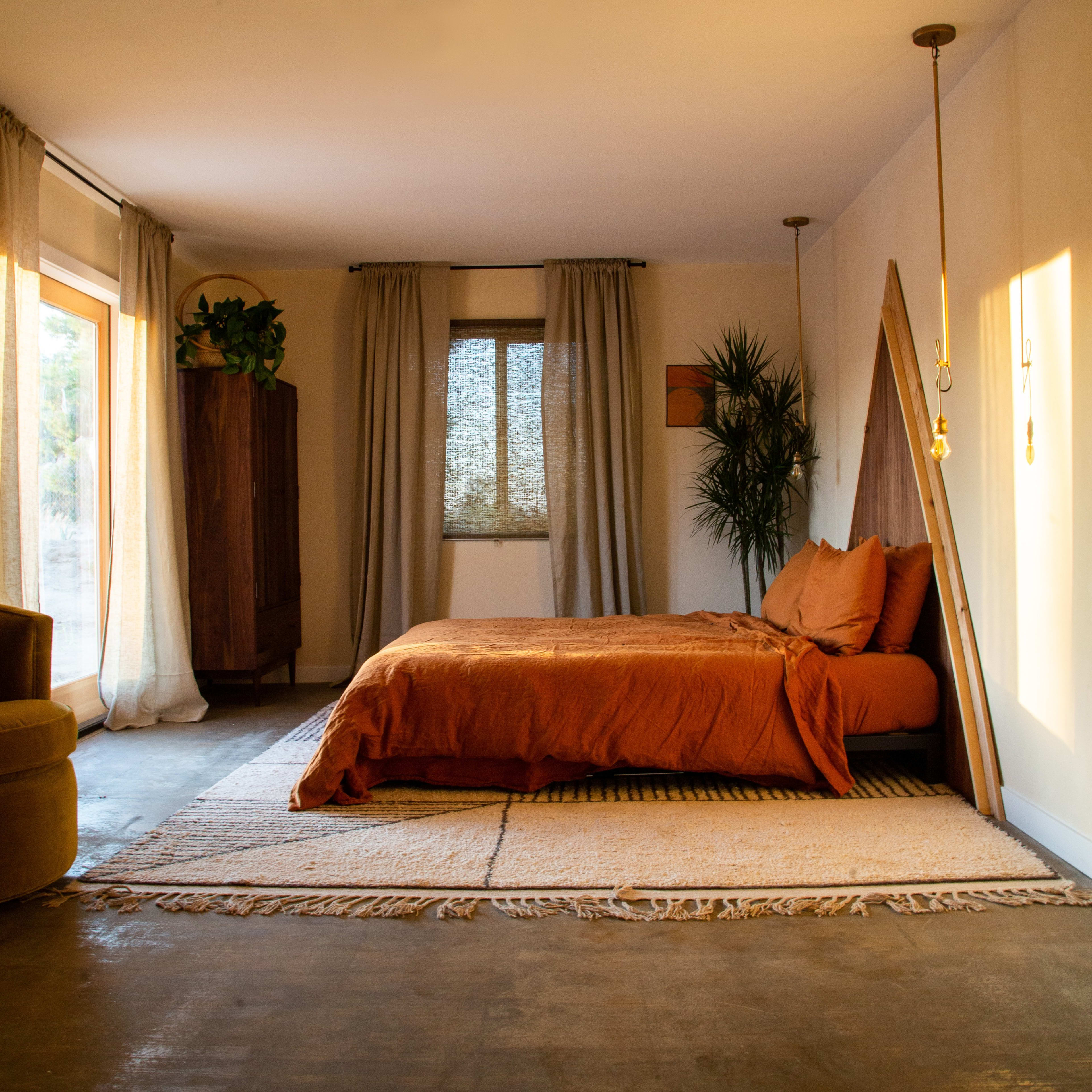 A spacious bedroom featuring an orange bedspread, large windows with sheer curtains, and a potted plant beside the bed.