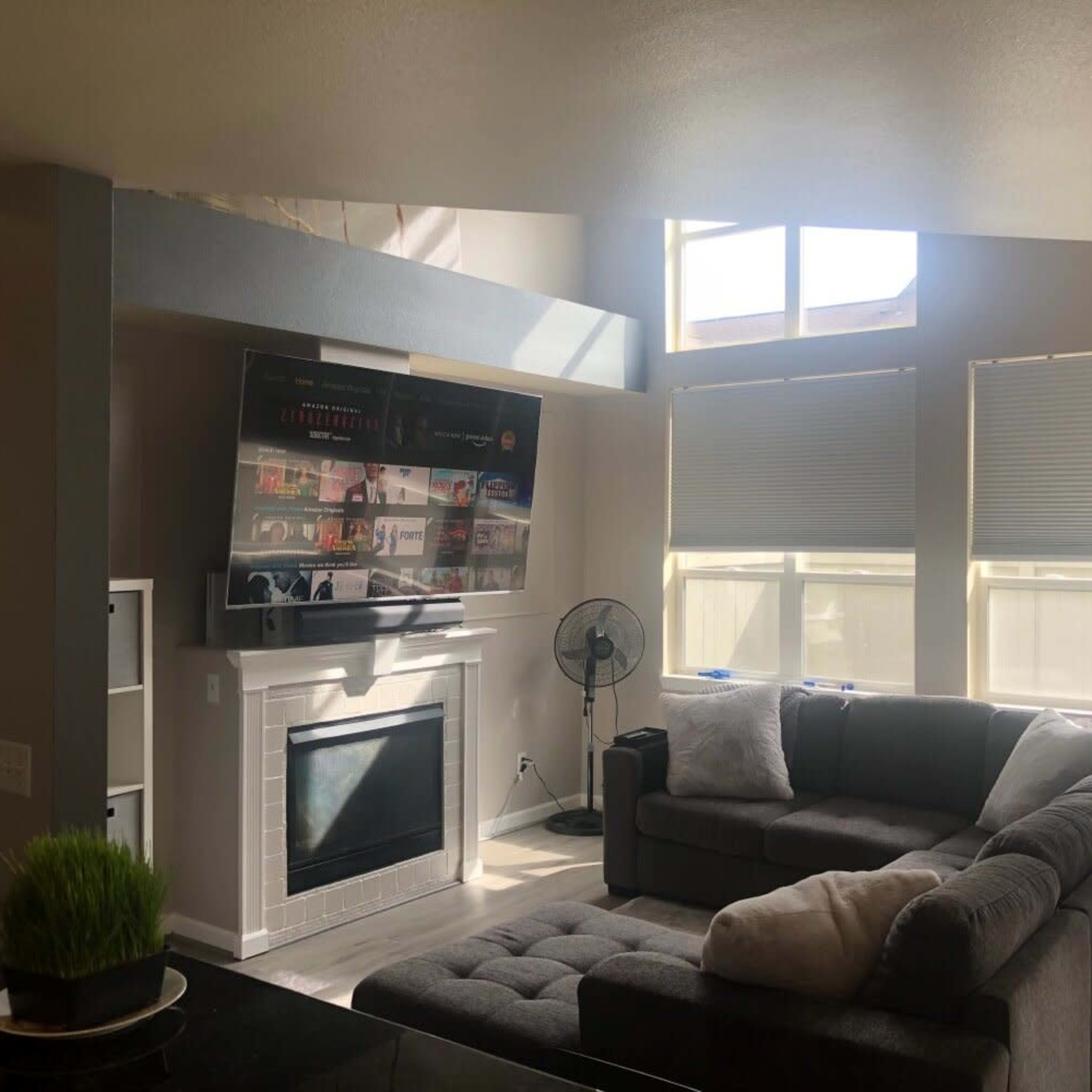 A modern living room with a large gray sectional sofa, a television mounted above a fireplace, and bright windows allowing natural light to enter the space.