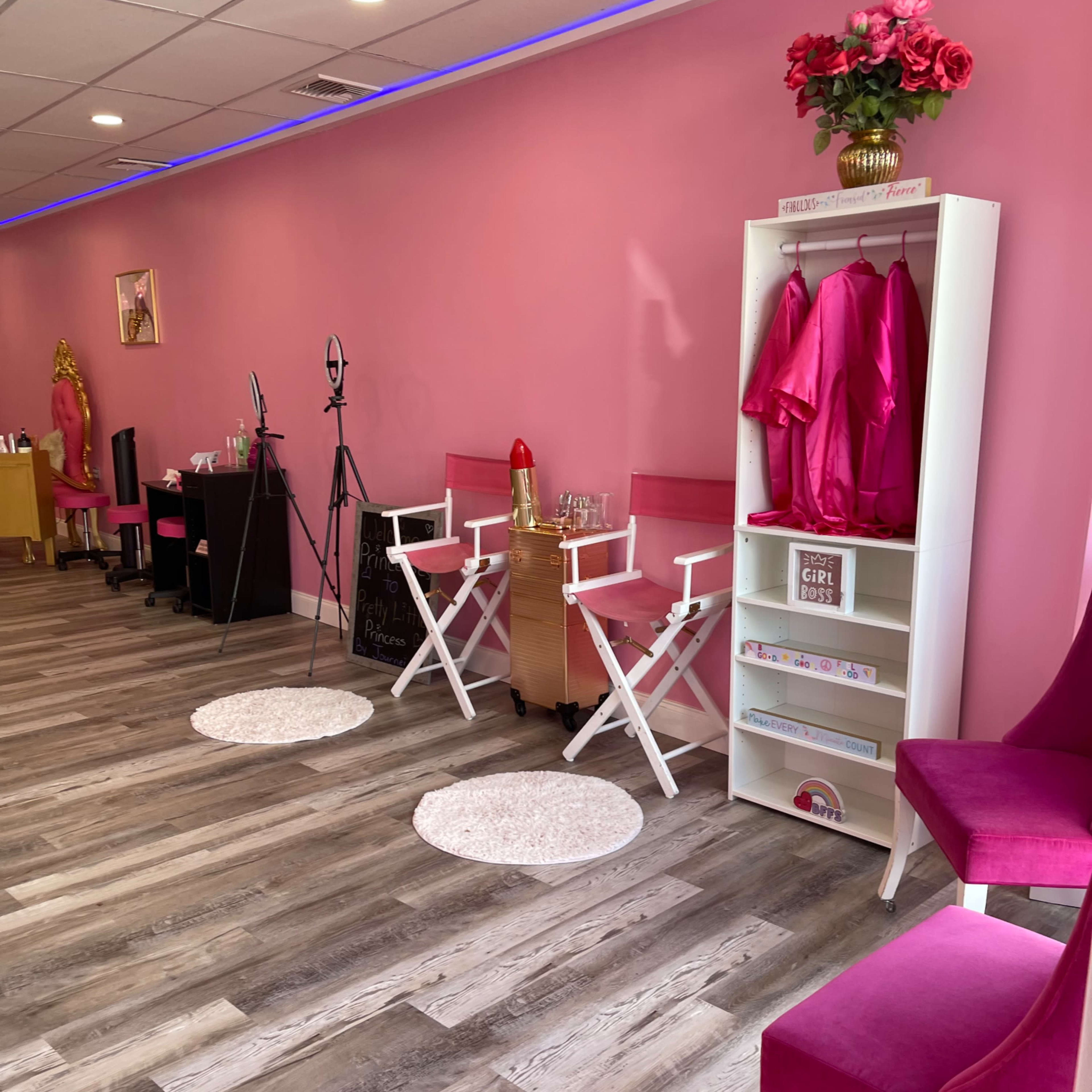 A brightly lit interior of a beauty studio features pink walls, wooden flooring, and styling chairs alongside a rack with pink garments and decor.
