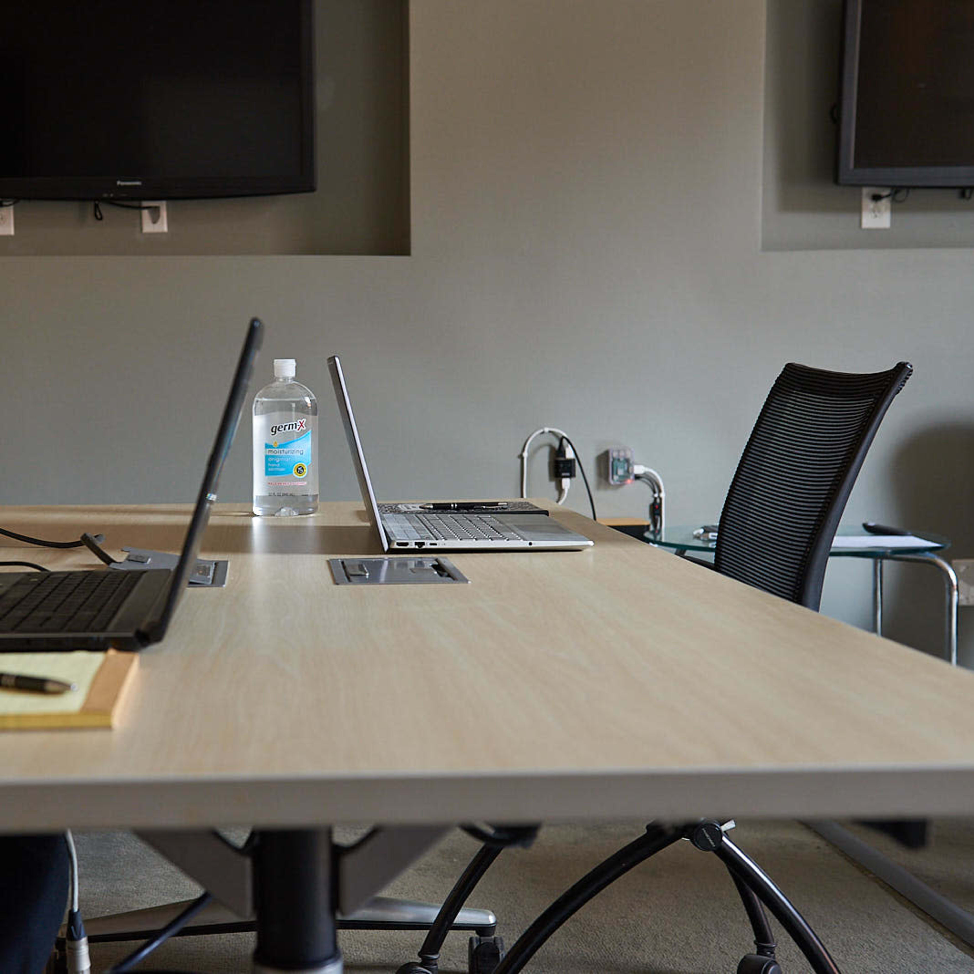 A conference room table is set up with two laptops, a notepad, a pen, and a bottle of water, with two black chairs positioned at the table.