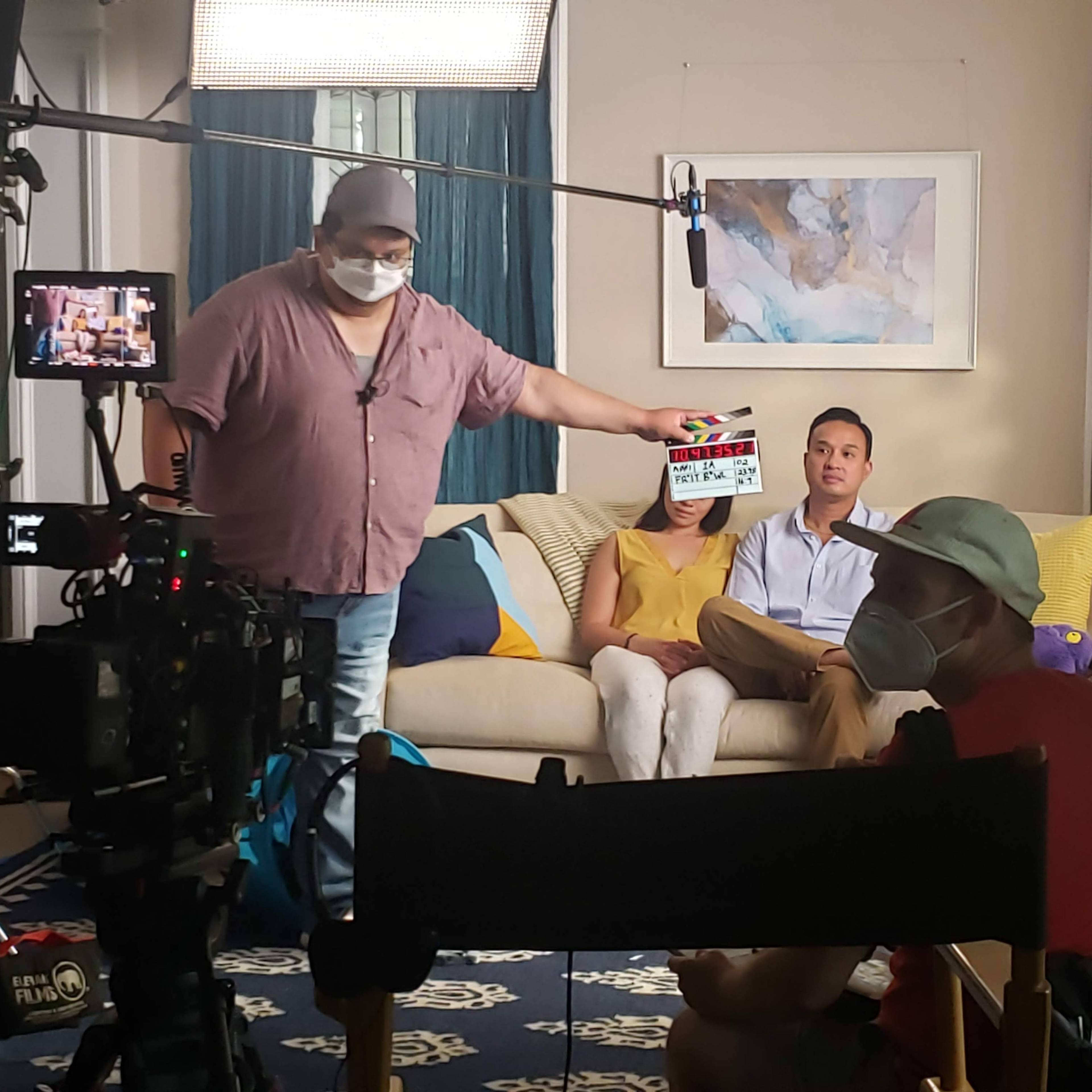 A film crew sets up a scene with a director holding a clapperboard in front of two actors sitting on a couch.
