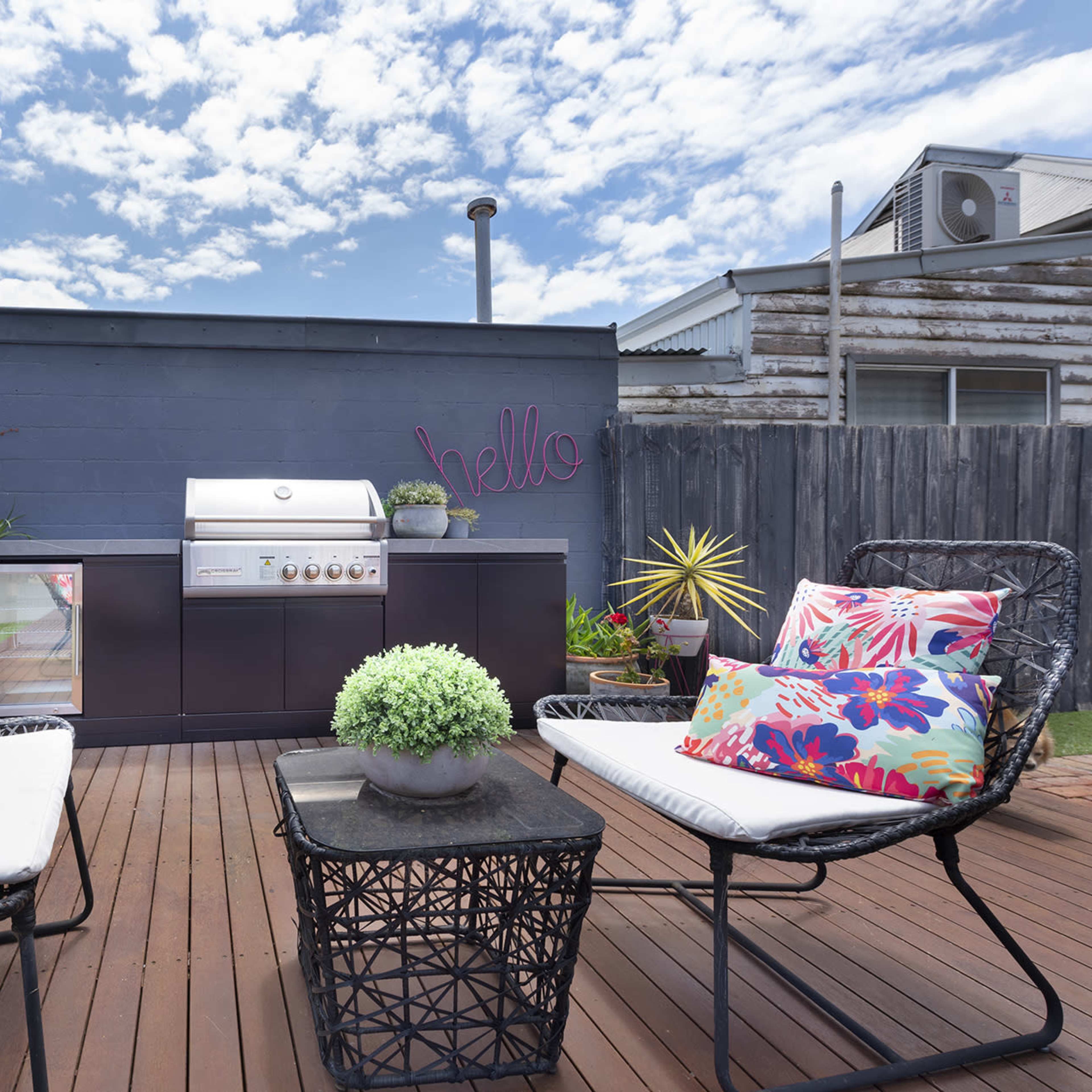 A backyard patio featuring a barbecue grill, potted plants, and two chairs with colorful cushions on a wooden deck.