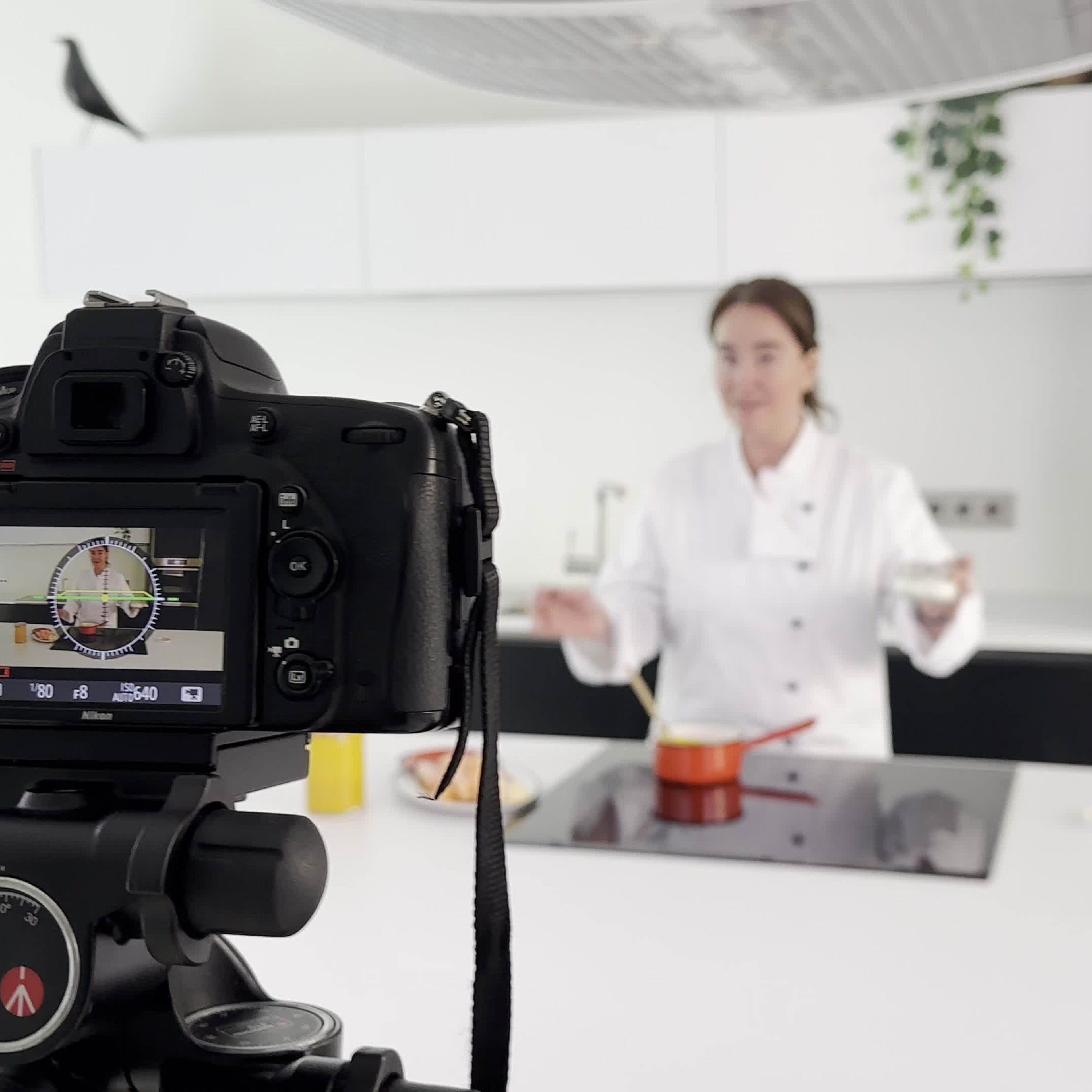 A camera on a tripod is focused on a woman in a chef's jacket gesturing while standing behind a kitchen island with a pot on the stove.