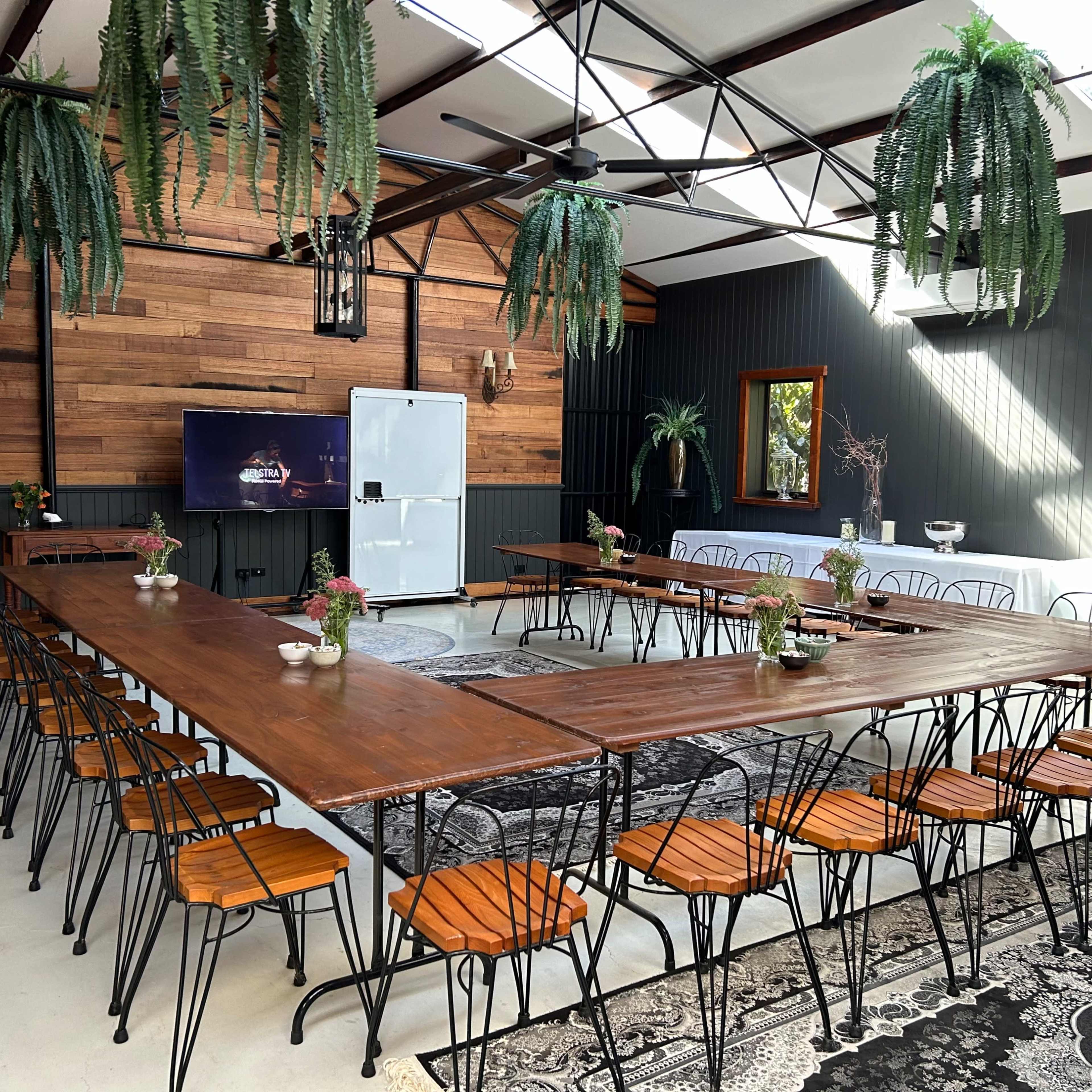 A long wooden table surrounded by metal chairs fills a bright, airy meeting room with greenery and decorative elements.