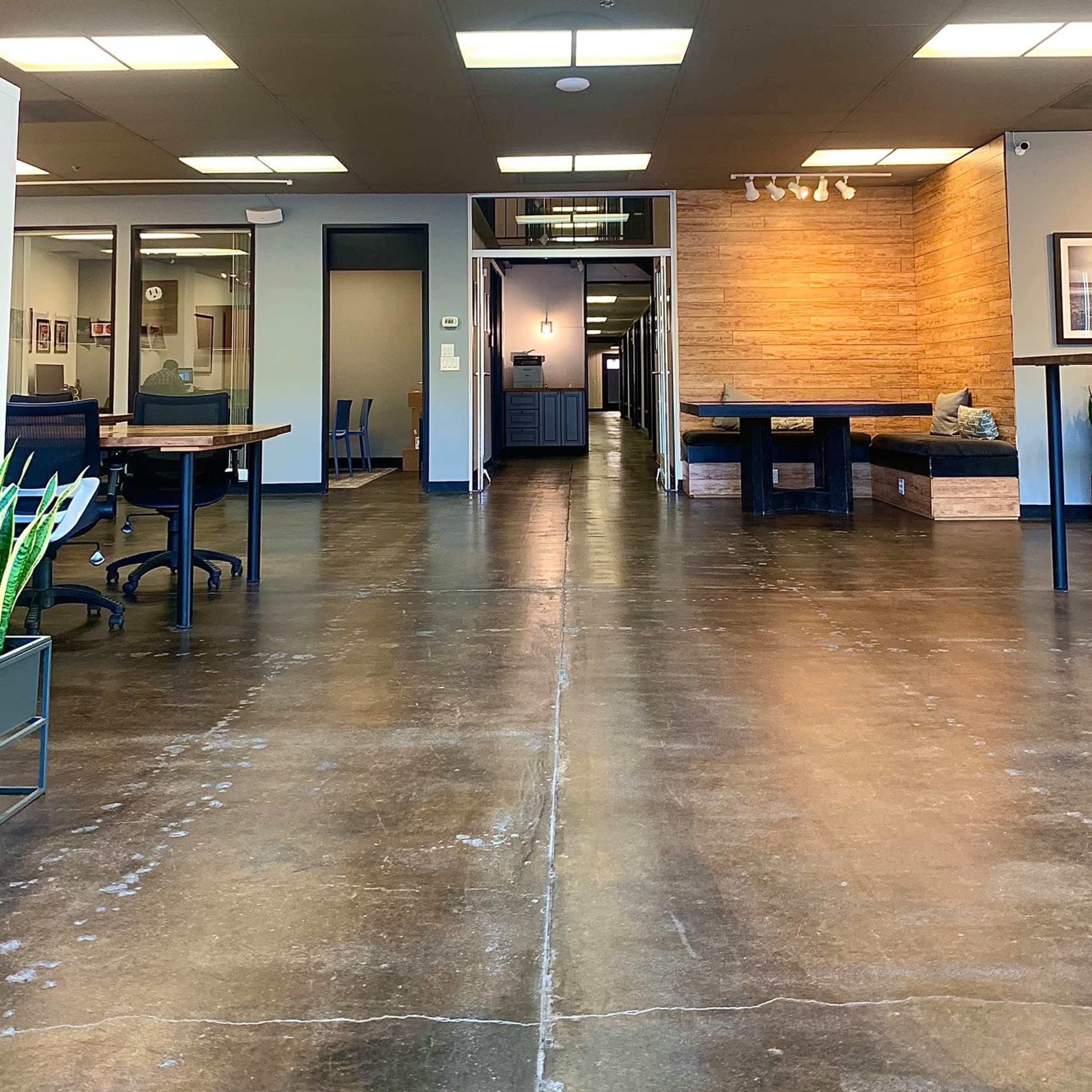 An open office space with a polished concrete floor, featuring several workstations on the left and a hallway leading to a wooden-paneled area in the background.