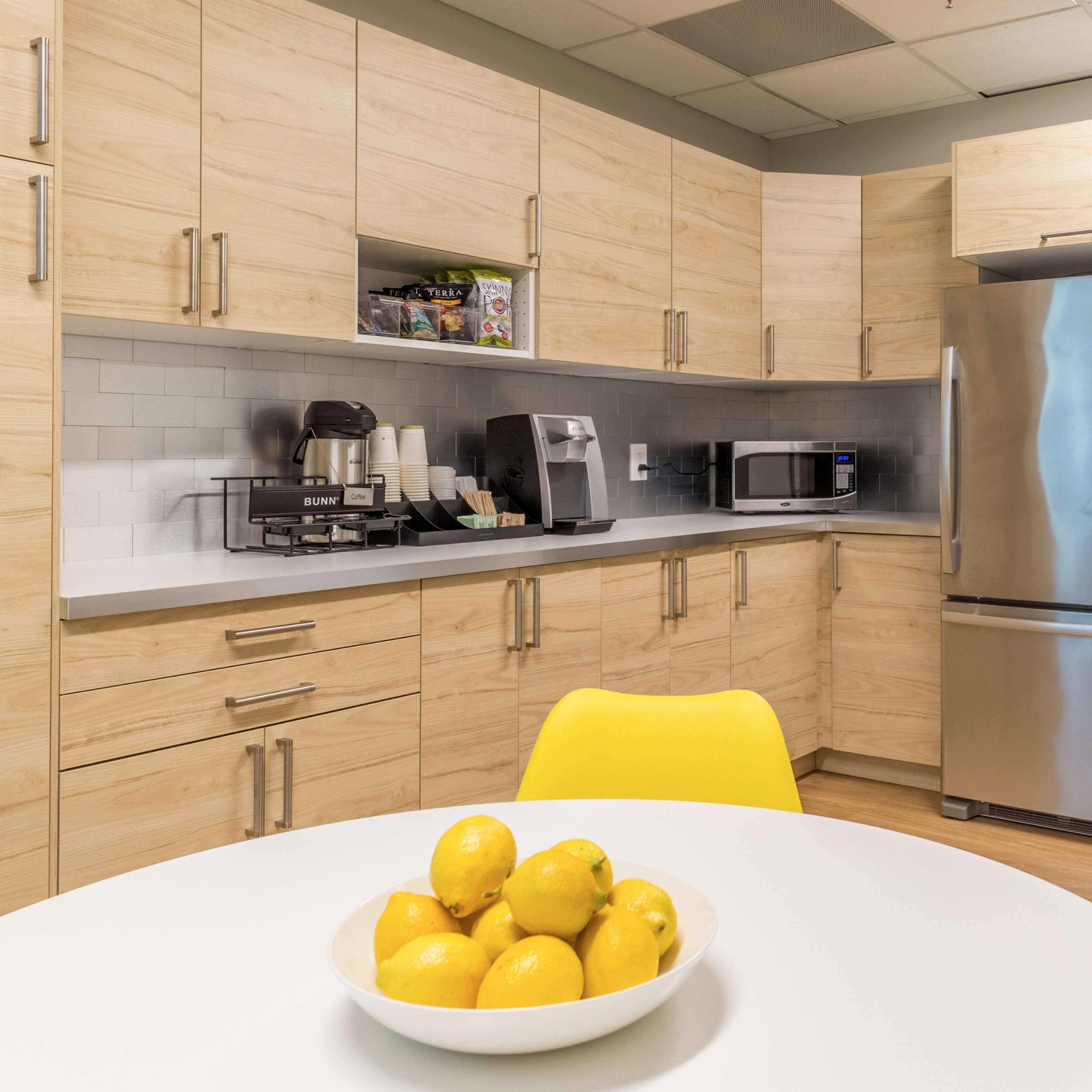 A modern kitchen features light wooden cabinets, stainless steel appliances, and a round dining table with a bowl of lemons.