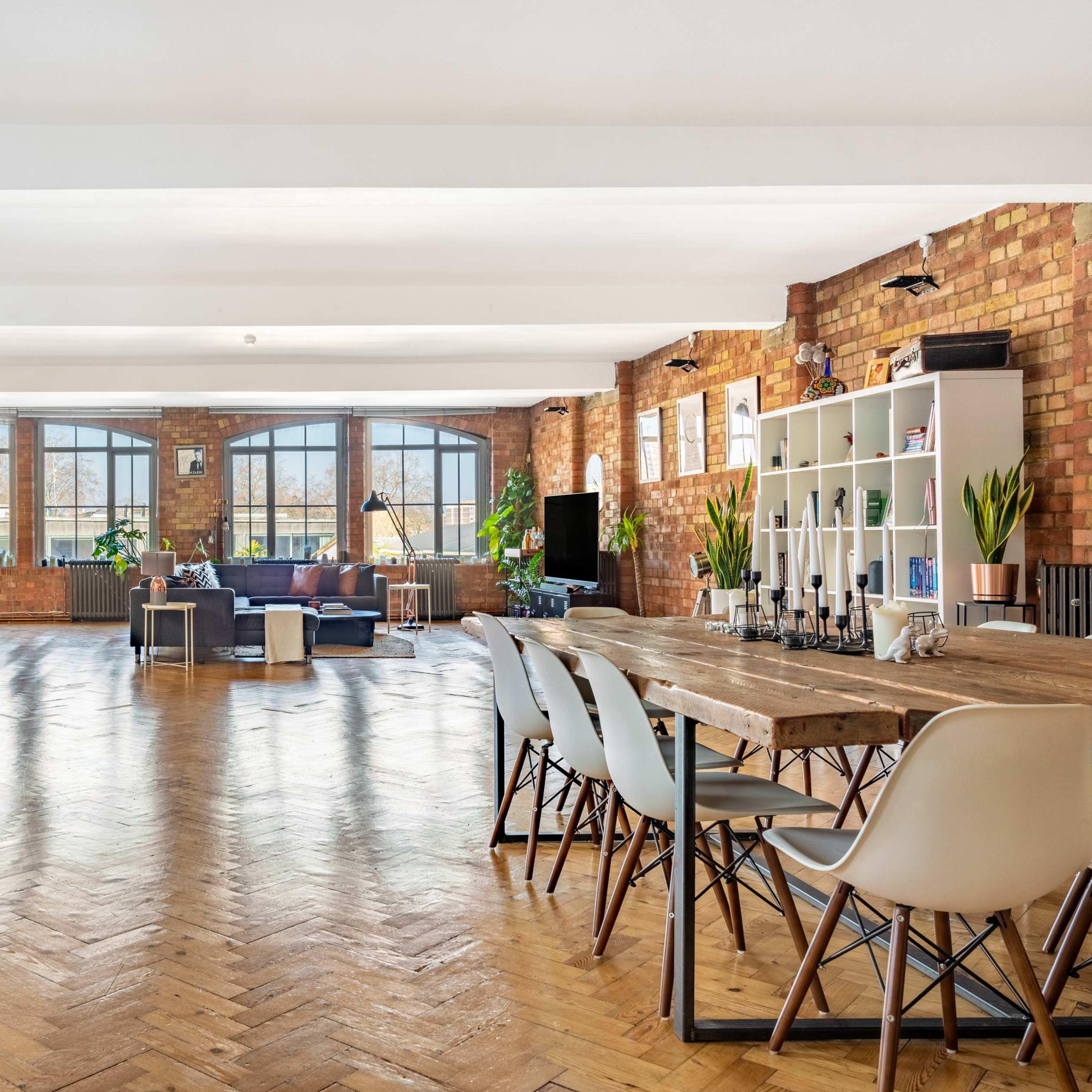 The spacious interior features exposed brick walls, large windows, and a mixture of modern furnishings and rustic decor, with a dining table in the foreground.