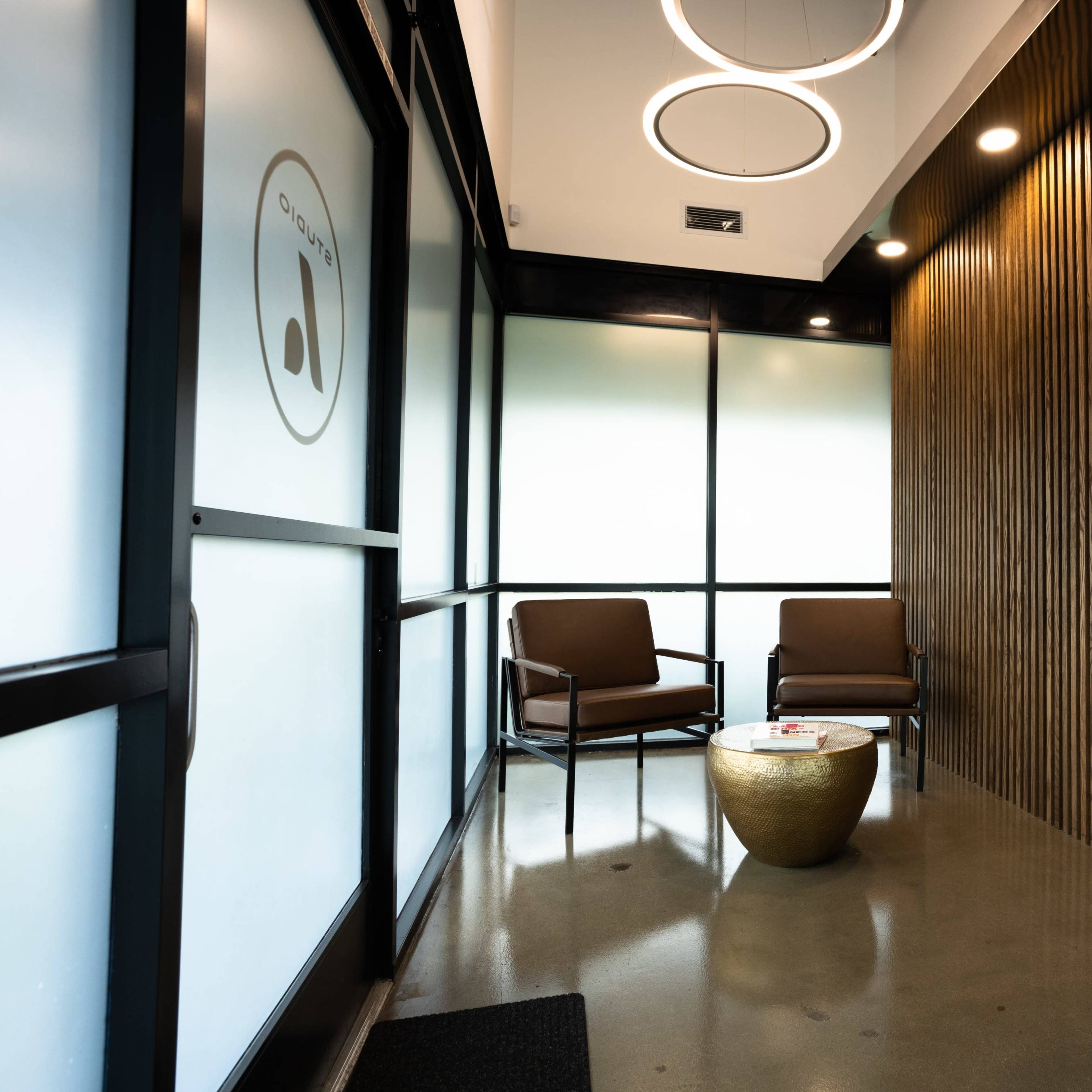 A modern waiting area featuring two brown chairs, a round gold coffee table, and green foliage beside a woven basket in a space with wood-paneled walls and large windows.