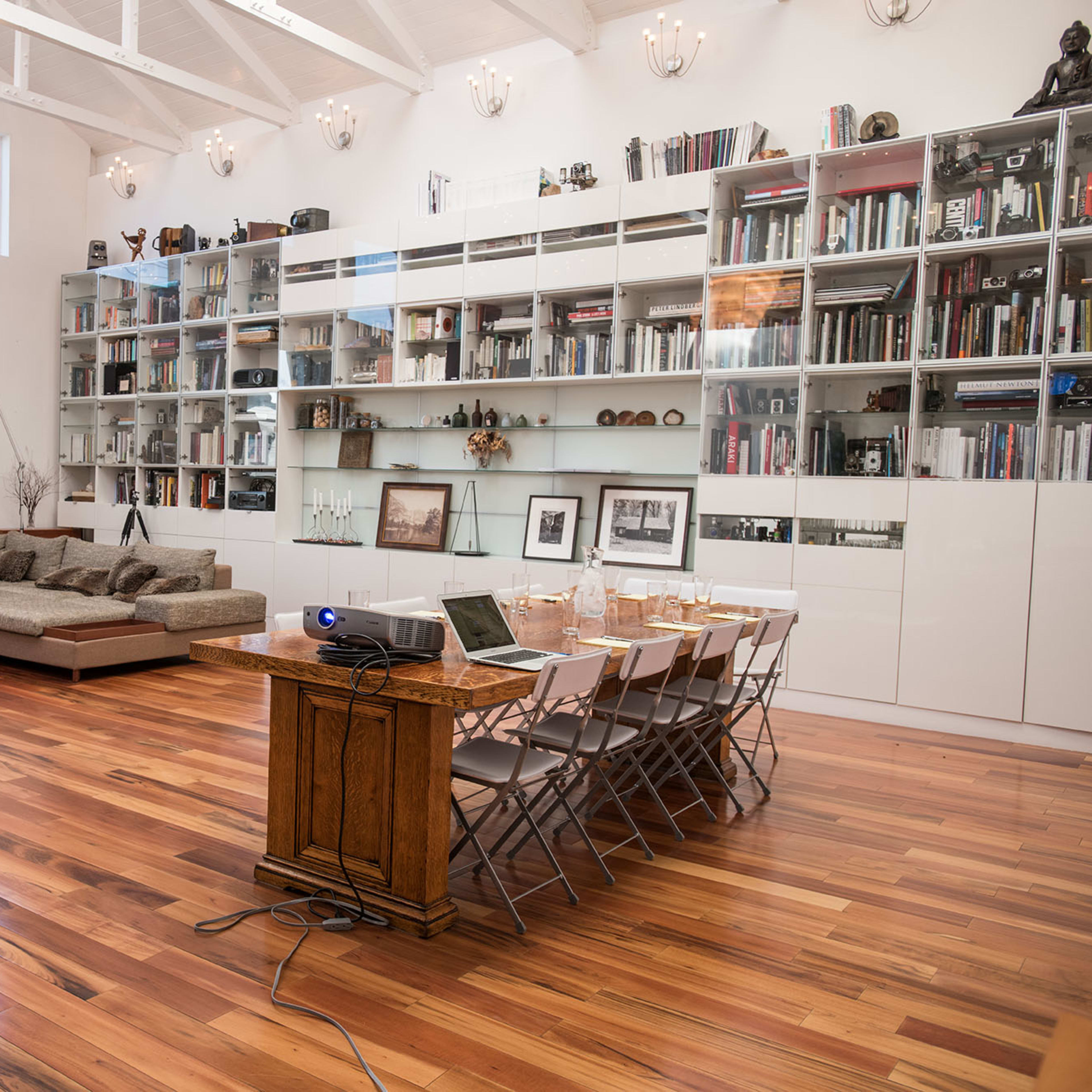 A modern meeting room with a wooden table set for a presentation, chairs arranged around it, and a large bookshelf filled with books along one wall.