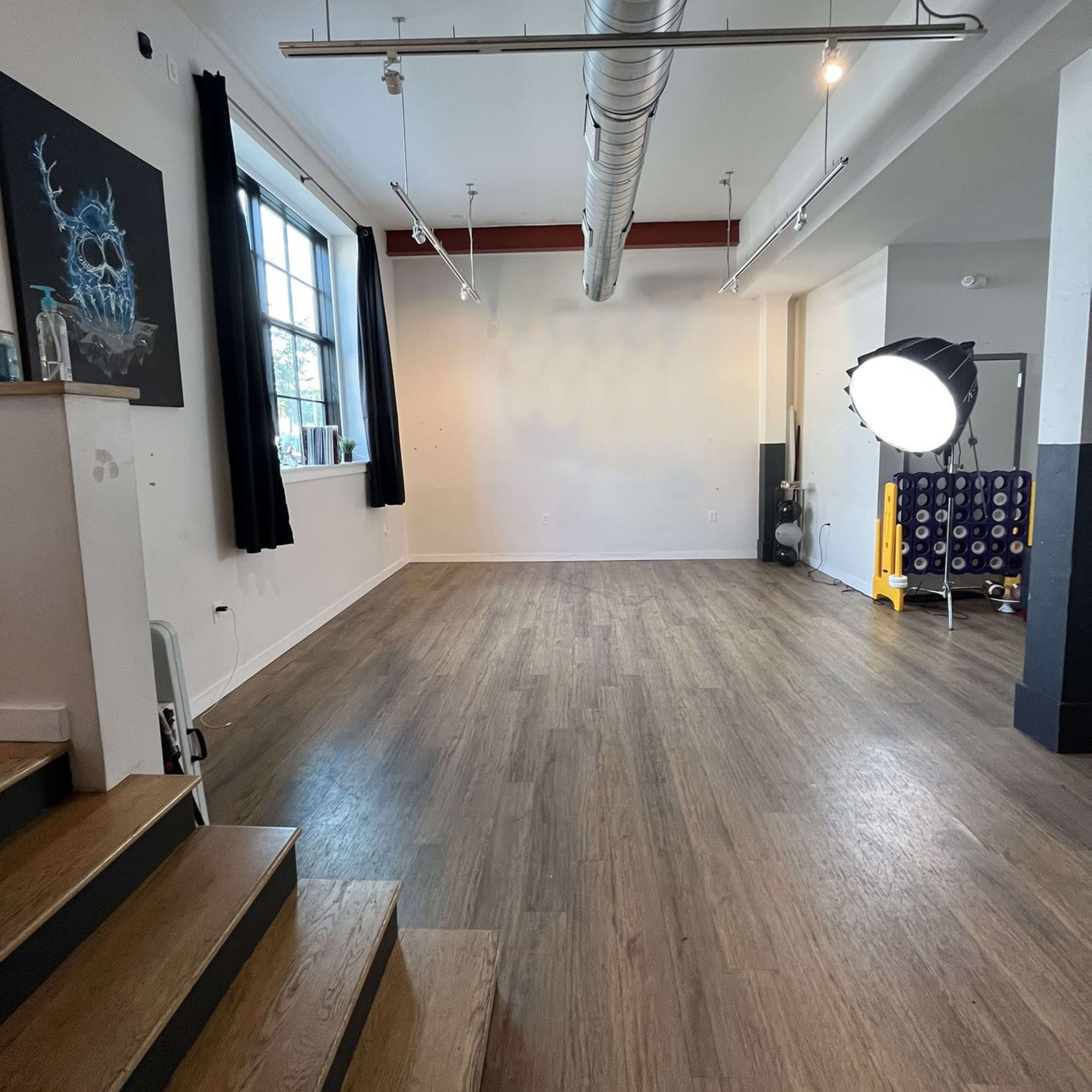 A spacious, empty studio with wooden flooring, large windows, and a spotlight set up in the corner.