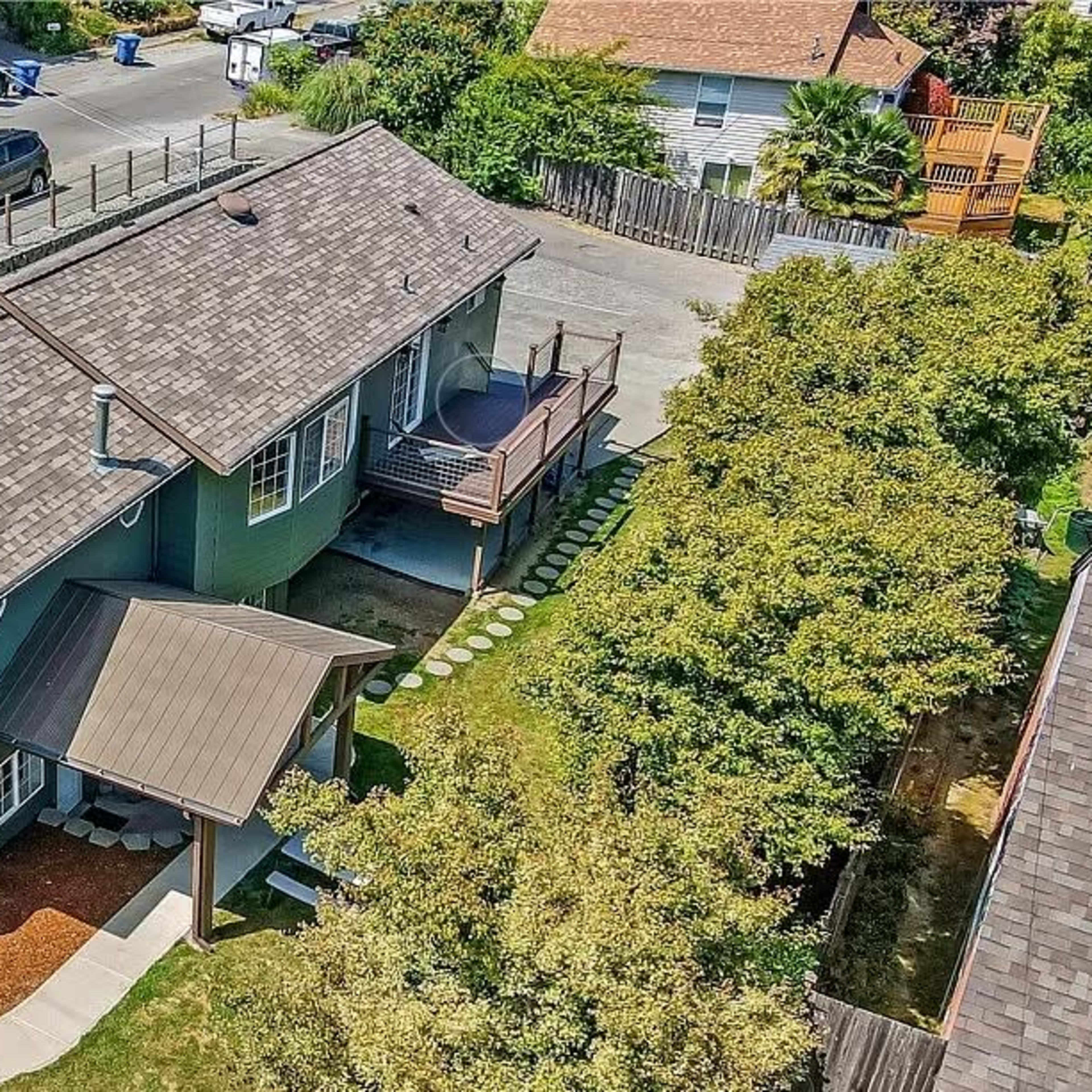 A green house with a sloped roof is situated on a corner lot surrounded by a fence, with parked cars visible along the street.
