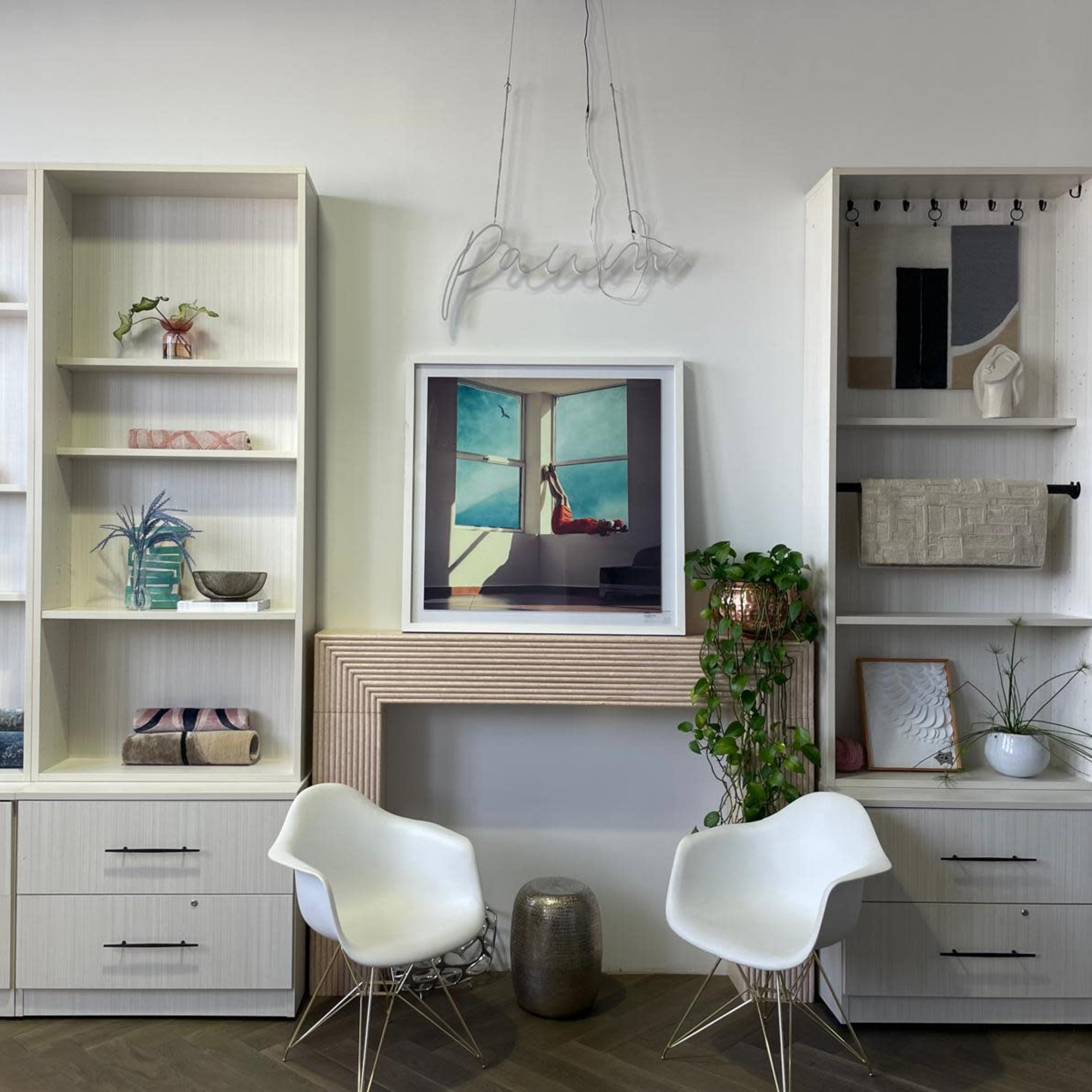 A modern living space features a light-colored shelving unit with decorative items, two white chairs, and a framed artwork above a streamlined fireplace.