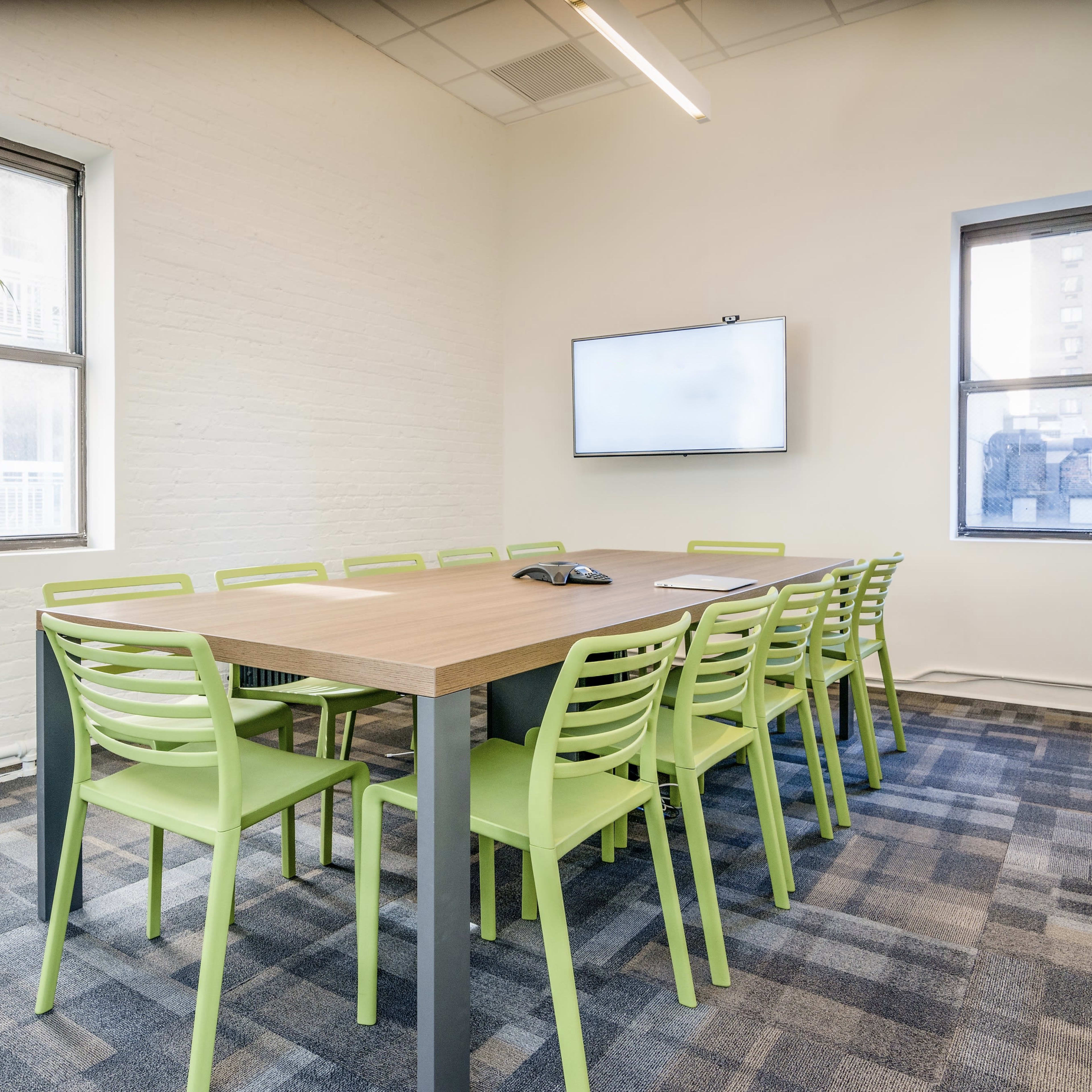 A meeting room features a long table surrounded by green chairs, with windows letting in natural light and a wall-mounted screen at one end.