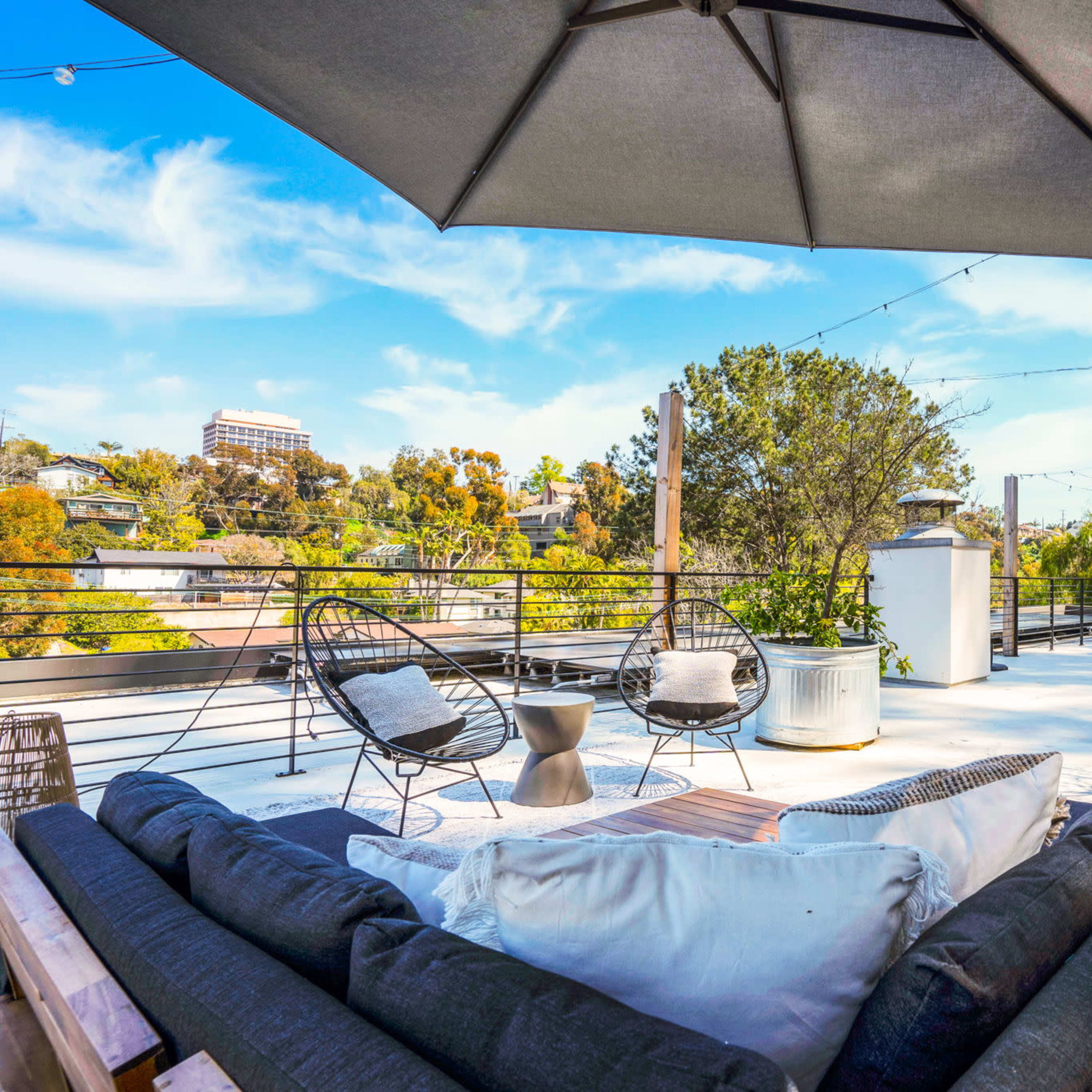 A rooftop patio featuring a sectional sofa, two chairs, an umbrella, and a view of surrounding greenery.
