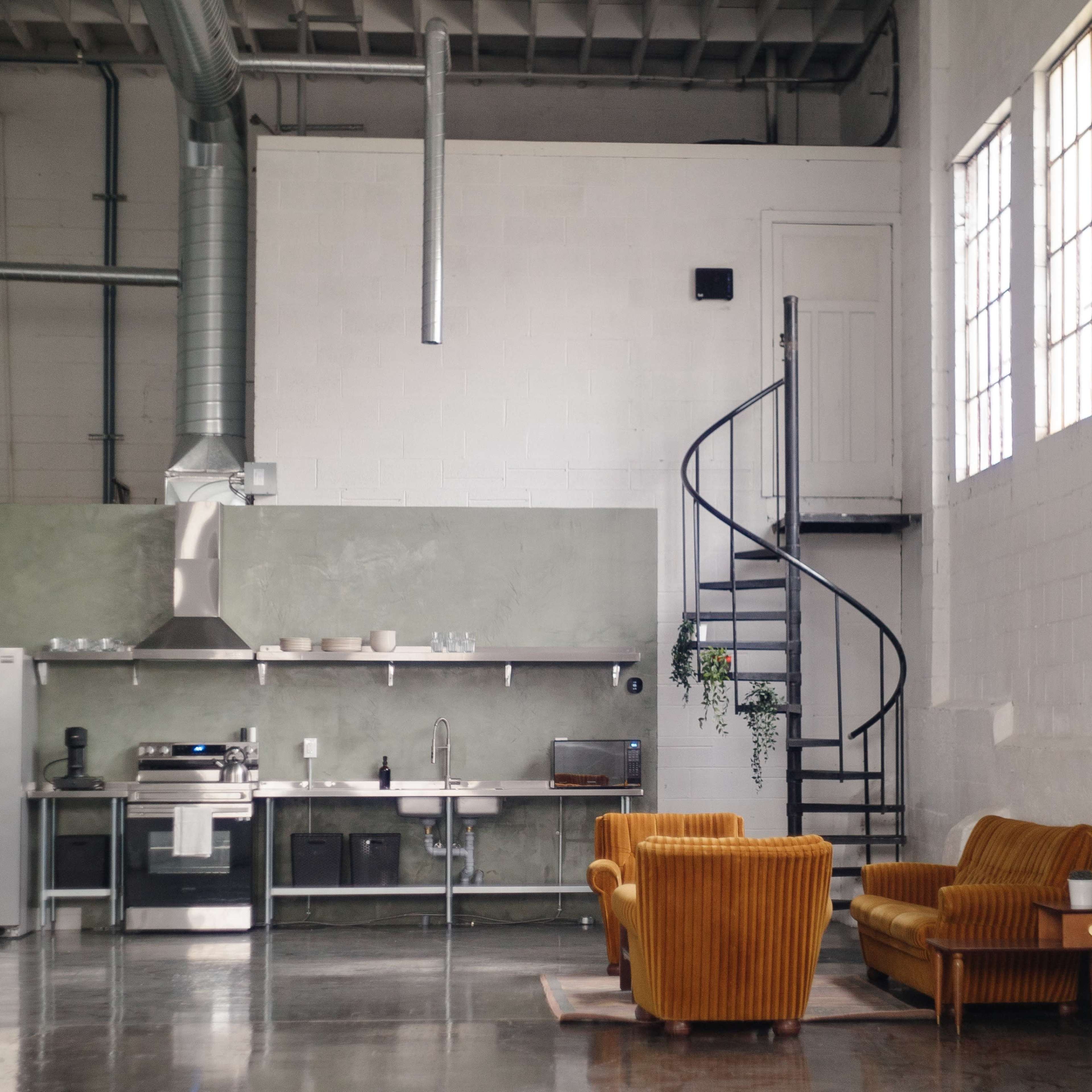 A modern kitchen with a spiral staircase, featuring two orange chairs, large windows, and industrial elements.