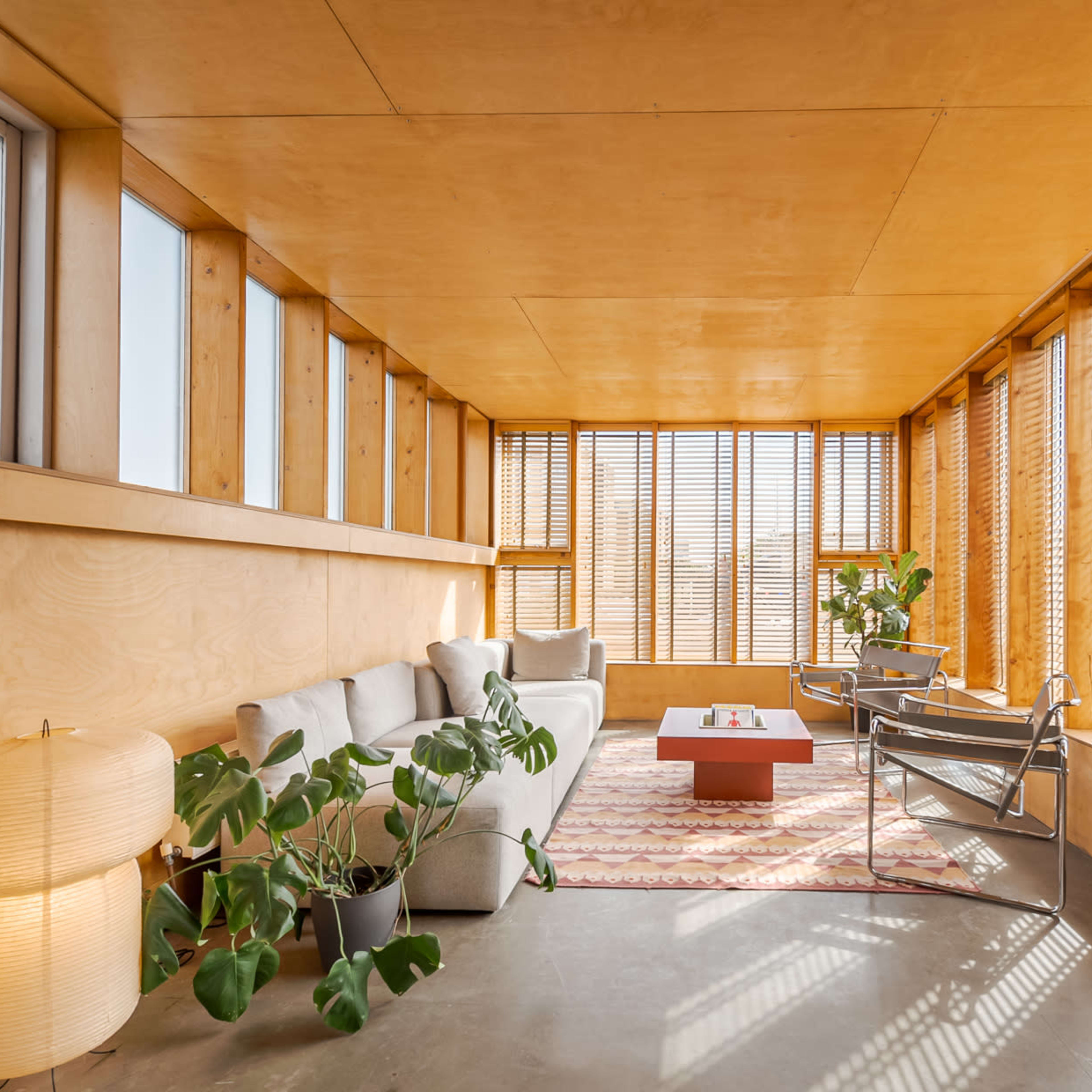 A modern, minimalistic living room with large windows, wooden walls, a light-colored sofa, a patterned rug, and potted plants.