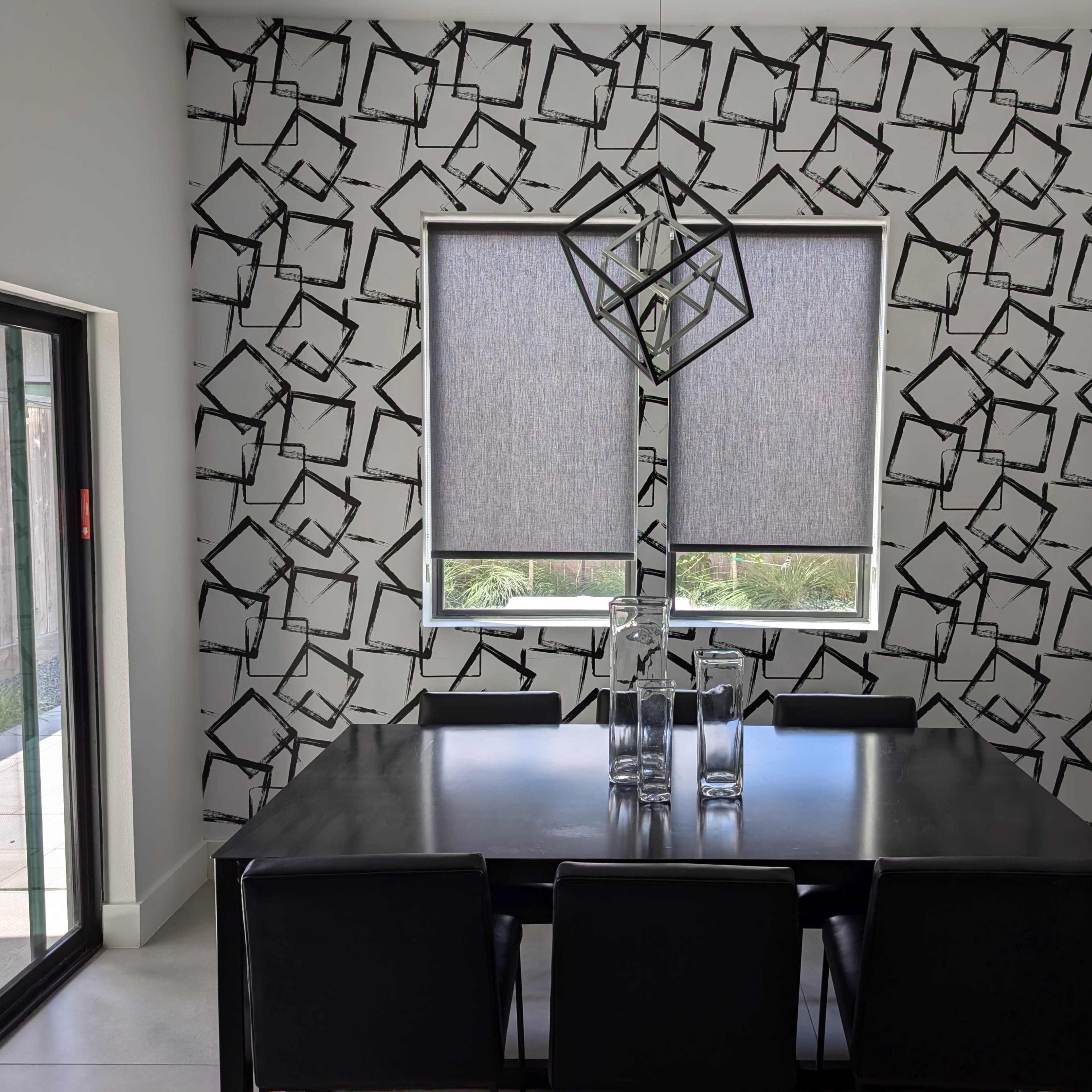 A dining area features a black and white patterned wall, a rectangular table with black chairs, and two large windows with roller shades.