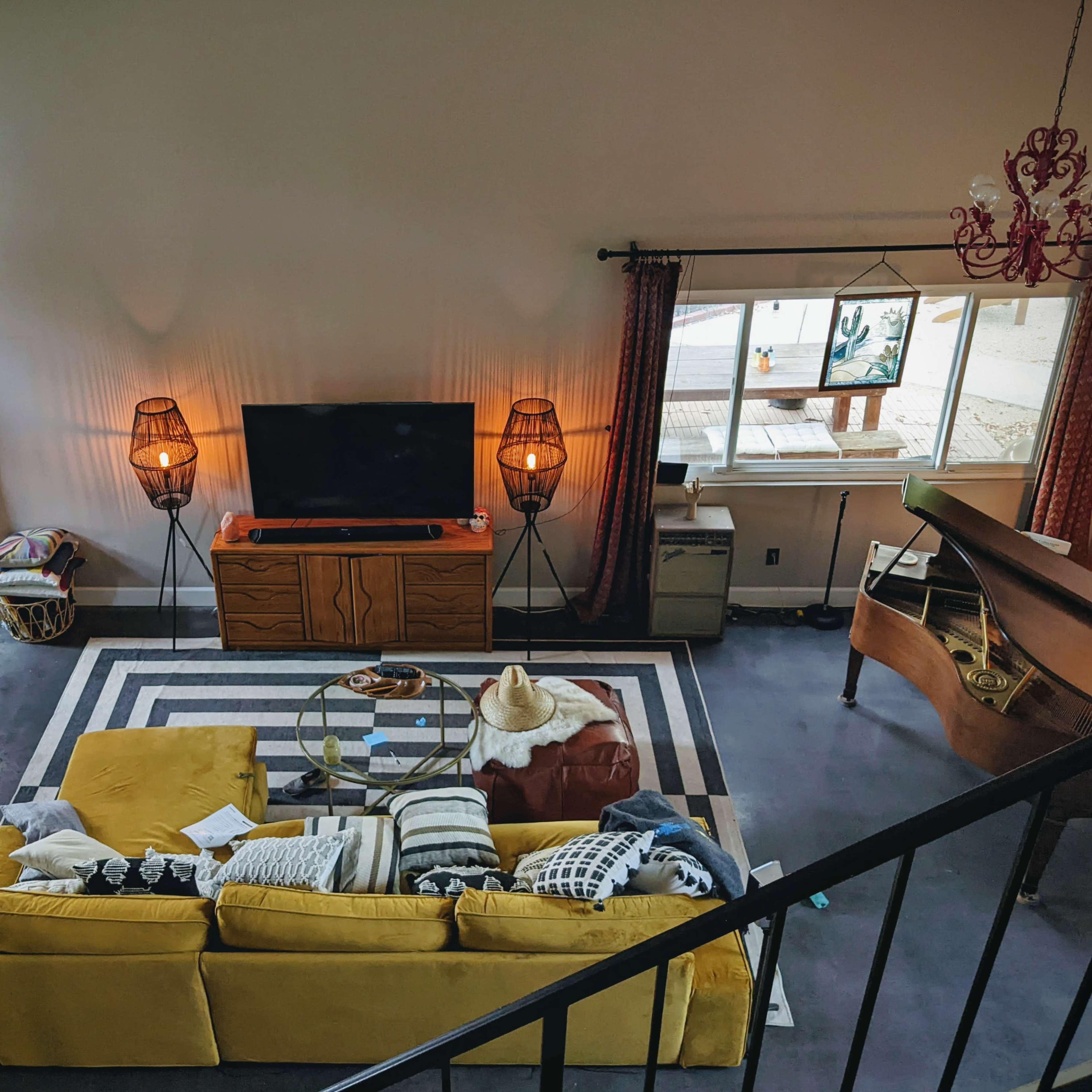 A spacious living room with a yellow sofa, a coffee table on a striped rug, a television on a wooden console, and large windows allowing natural light.