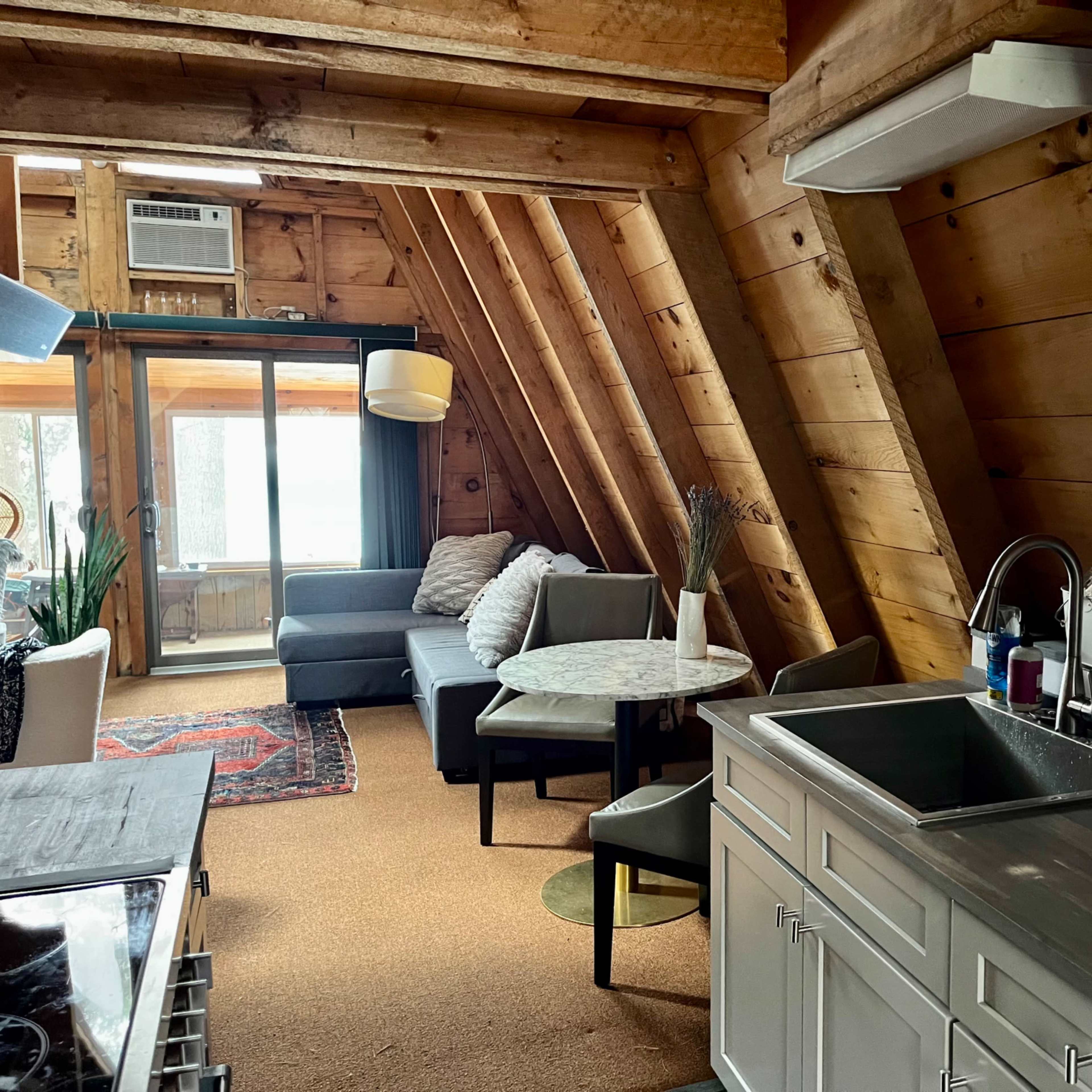 A cozy, wood-paneled cabin interior featuring a kitchen area with a sink, a dining table, and a living space with a sofa and large windows.