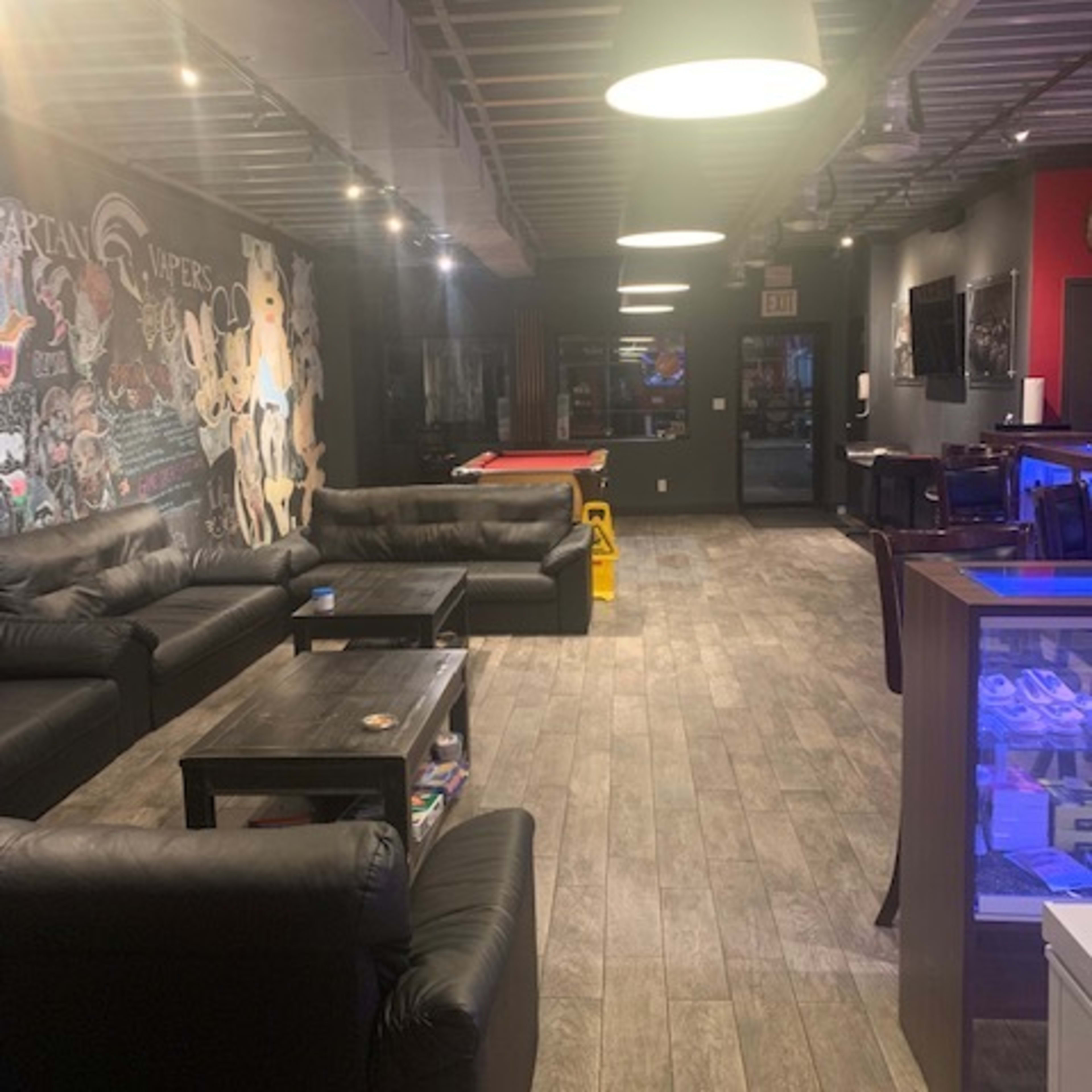 A dimly lit lounge area with black leather sofas, wooden tables, and a mural on the wall, with a pool table at the far end.