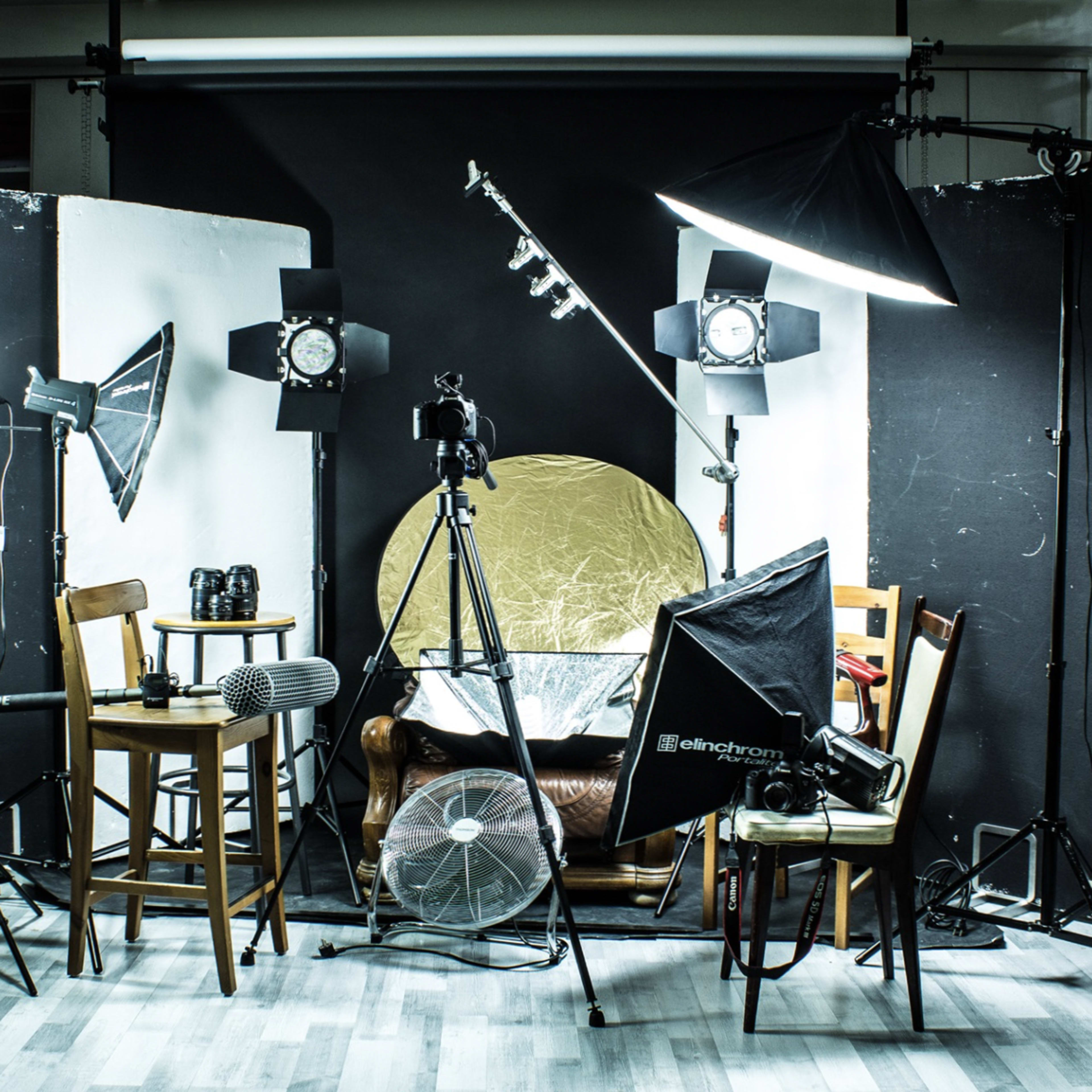 A photography studio setup with various lighting equipment, a camera on a tripod, and multiple chairs positioned around a backdrop.