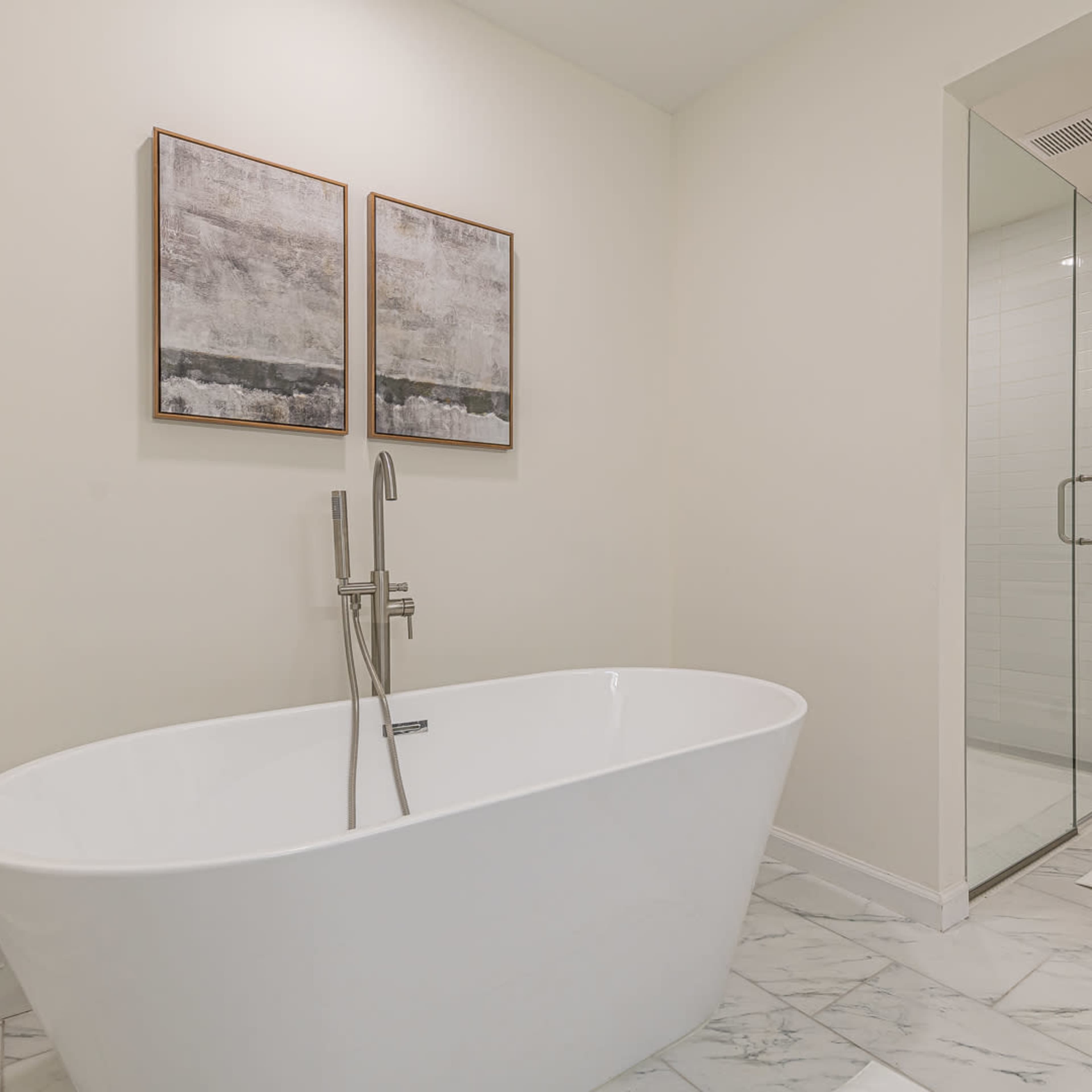 A modern bathroom featuring a freestanding white bathtub, two framed artwork pieces on the wall, and a glass shower enclosure.