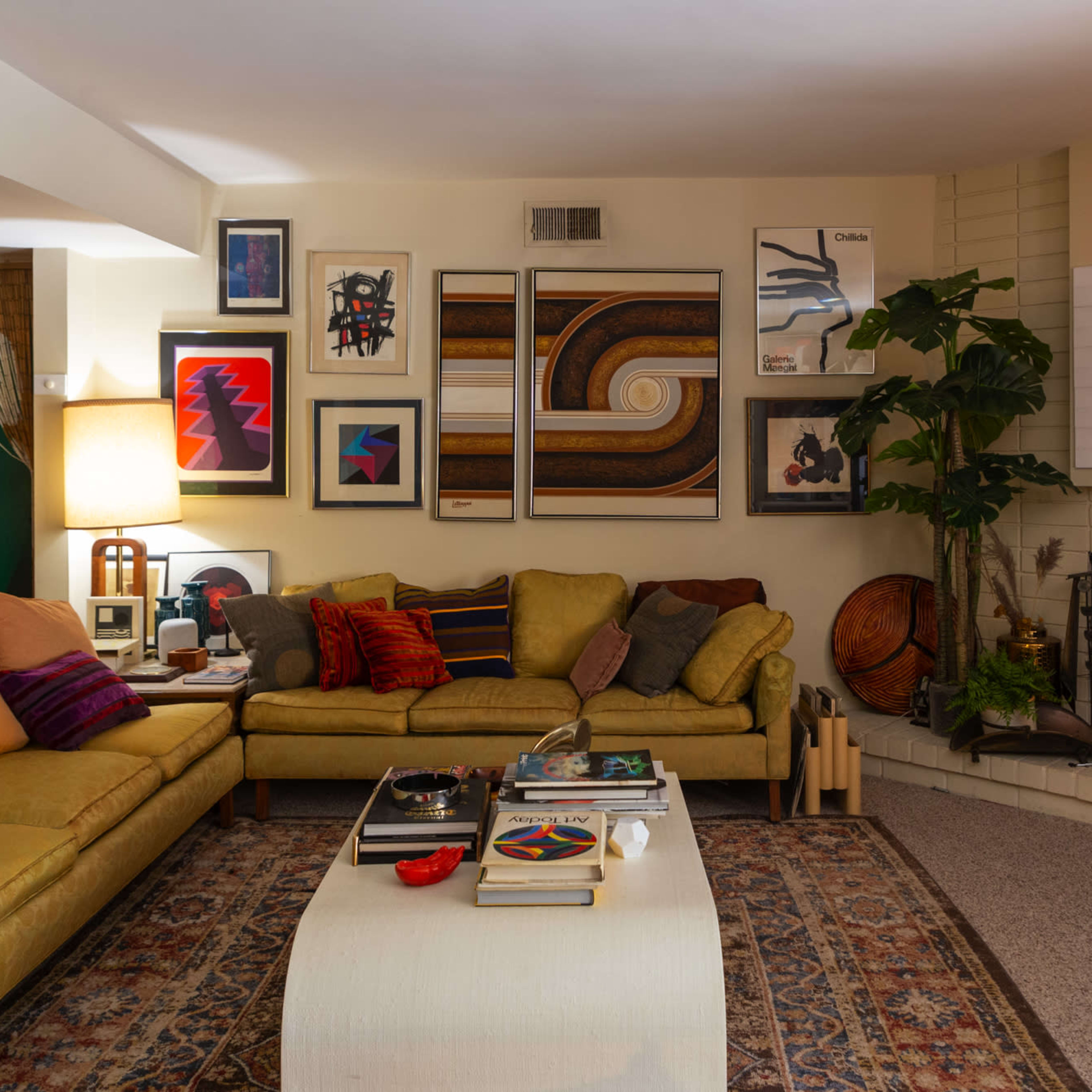 The living room features a green sectional sofa, a coffee table with various books and decor, a fireplace, and an arrangement of colorful artwork on the walls.