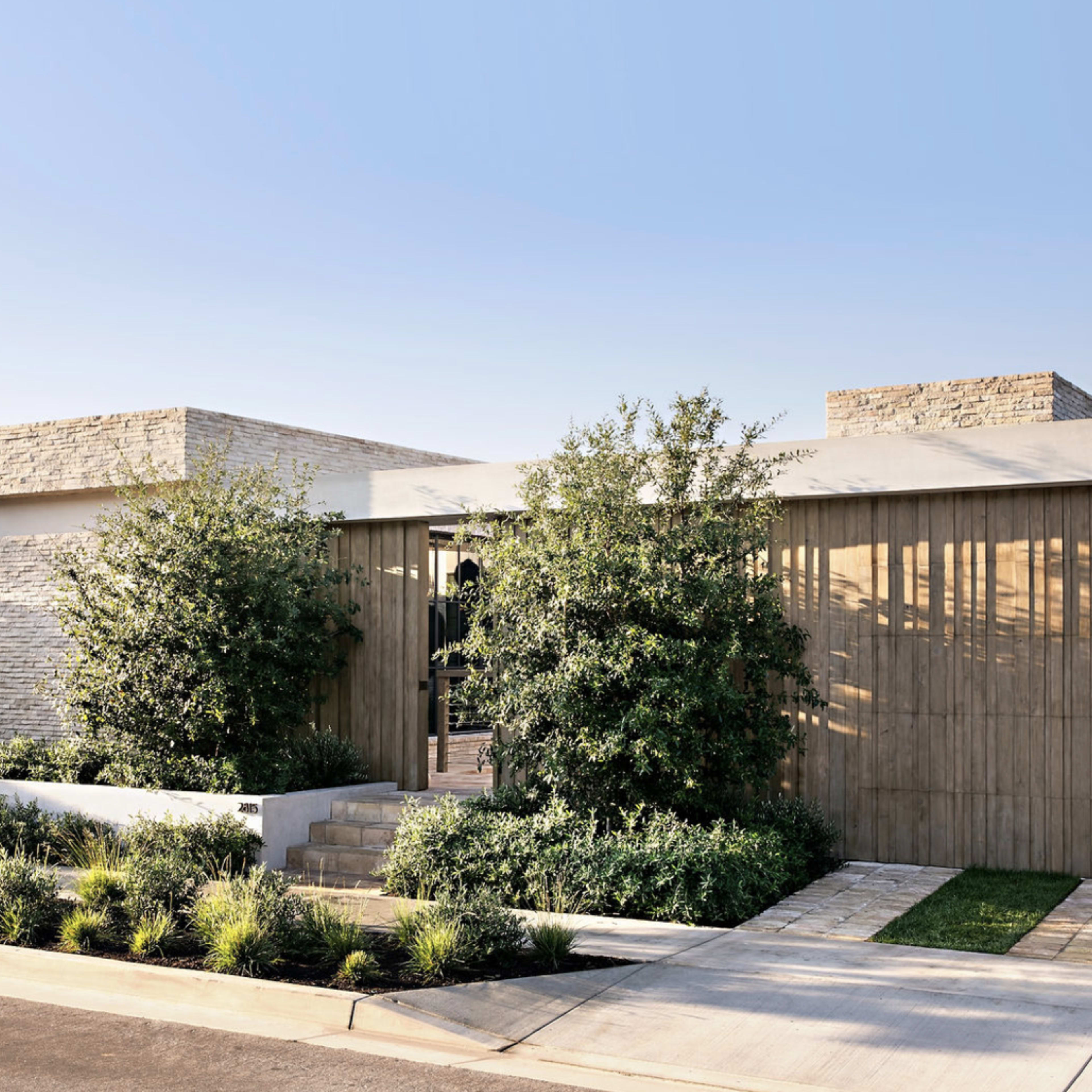 A modern house features a combination of stone and wood facade, surrounded by landscaped greenery and a pathway leading to the entrance.