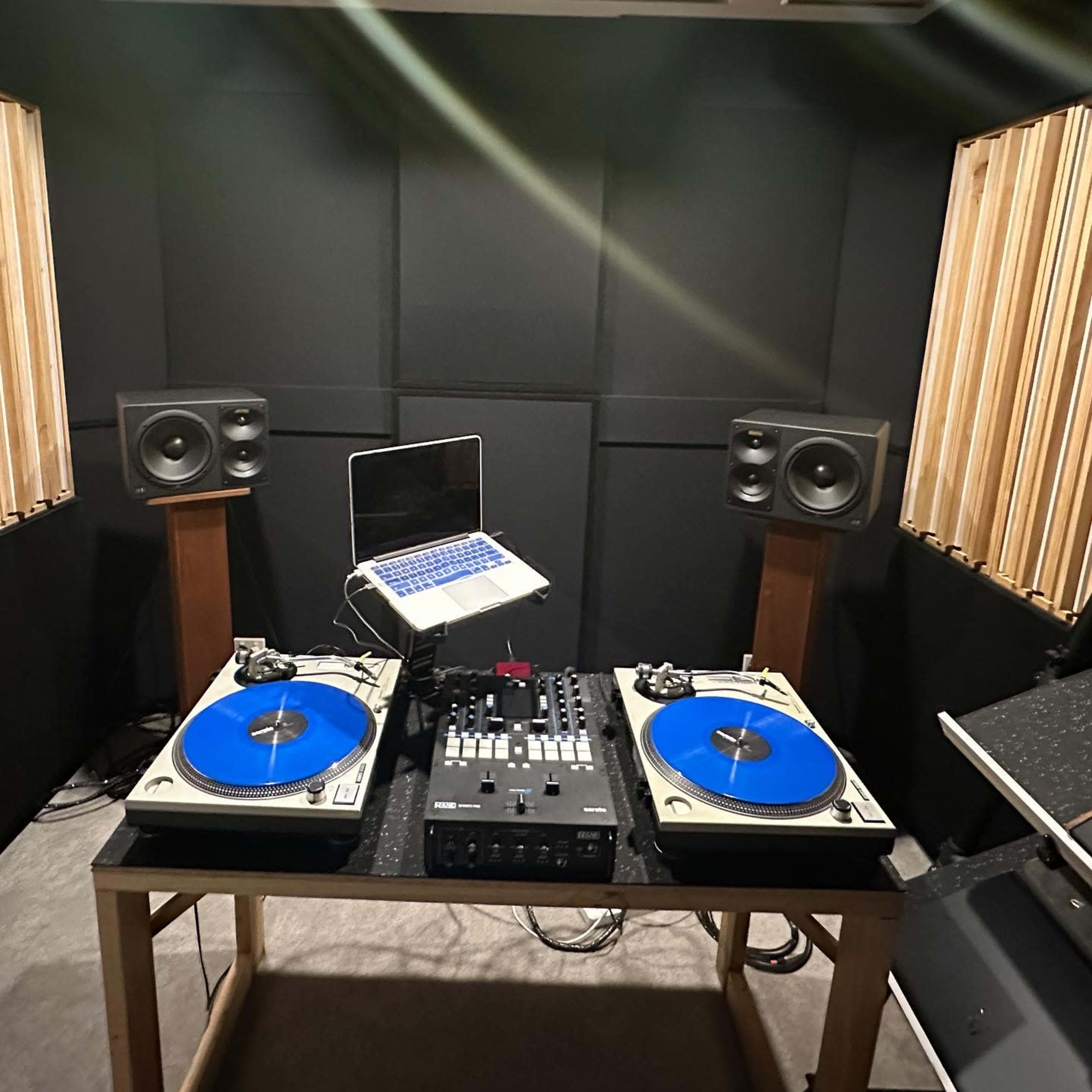 A DJ setup featuring two turntables with blue platters, a control mixer, and a laptop, all arranged in a soundproof room with wooden paneling.